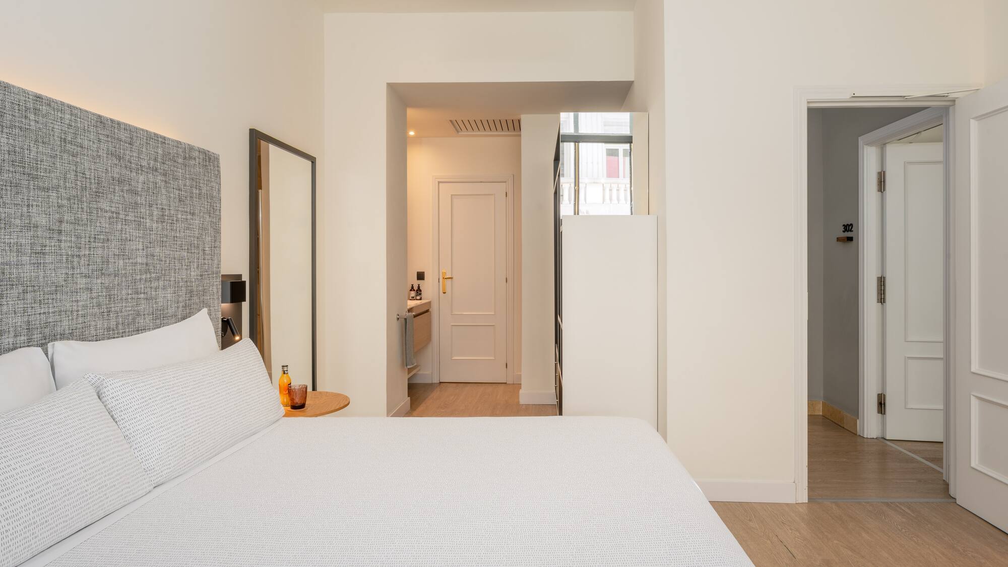 Modern hotel bedroom with white bed, grey headboard, and light wood floor.