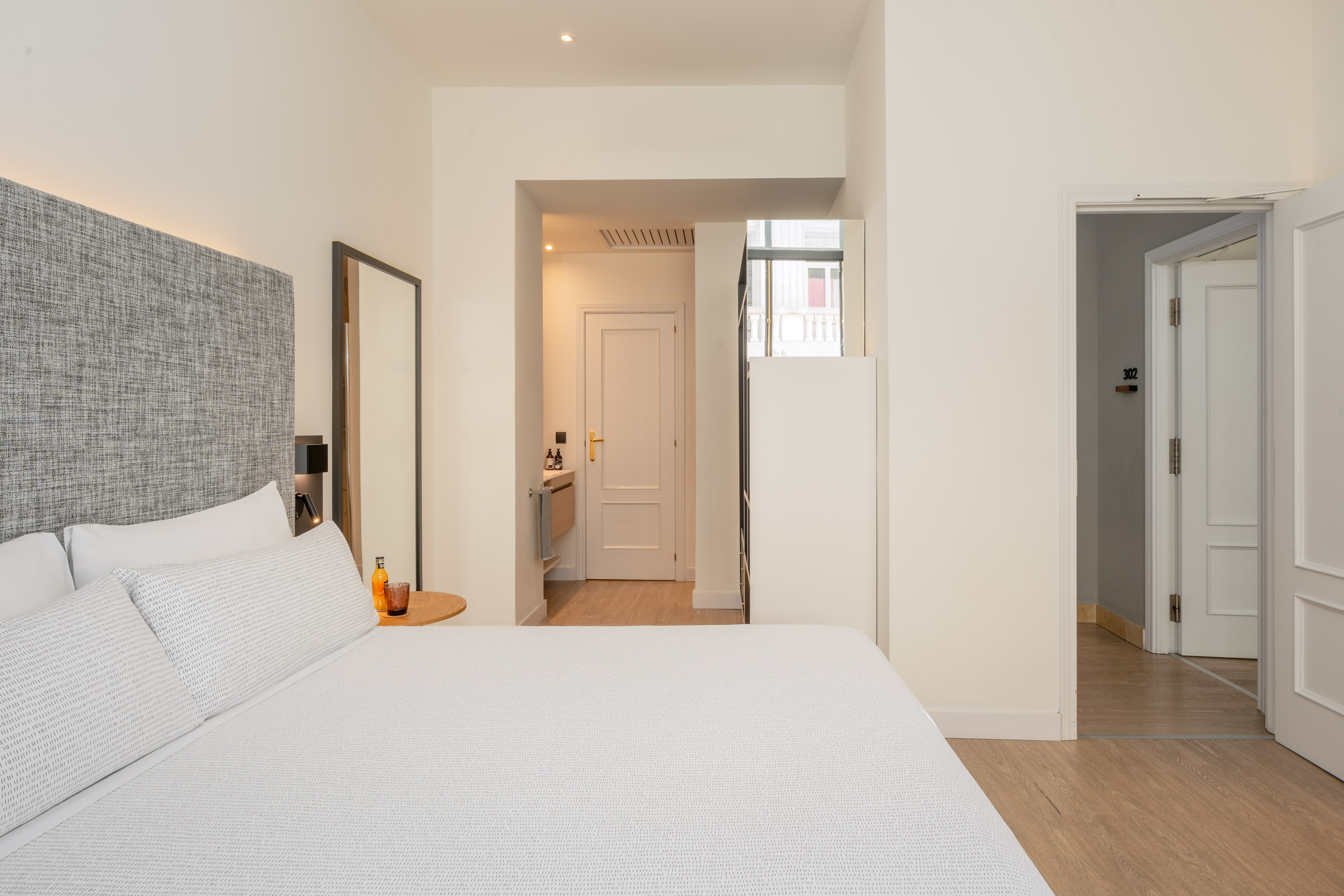 Modern hotel bedroom with white bed, grey headboard, and light wood floor.