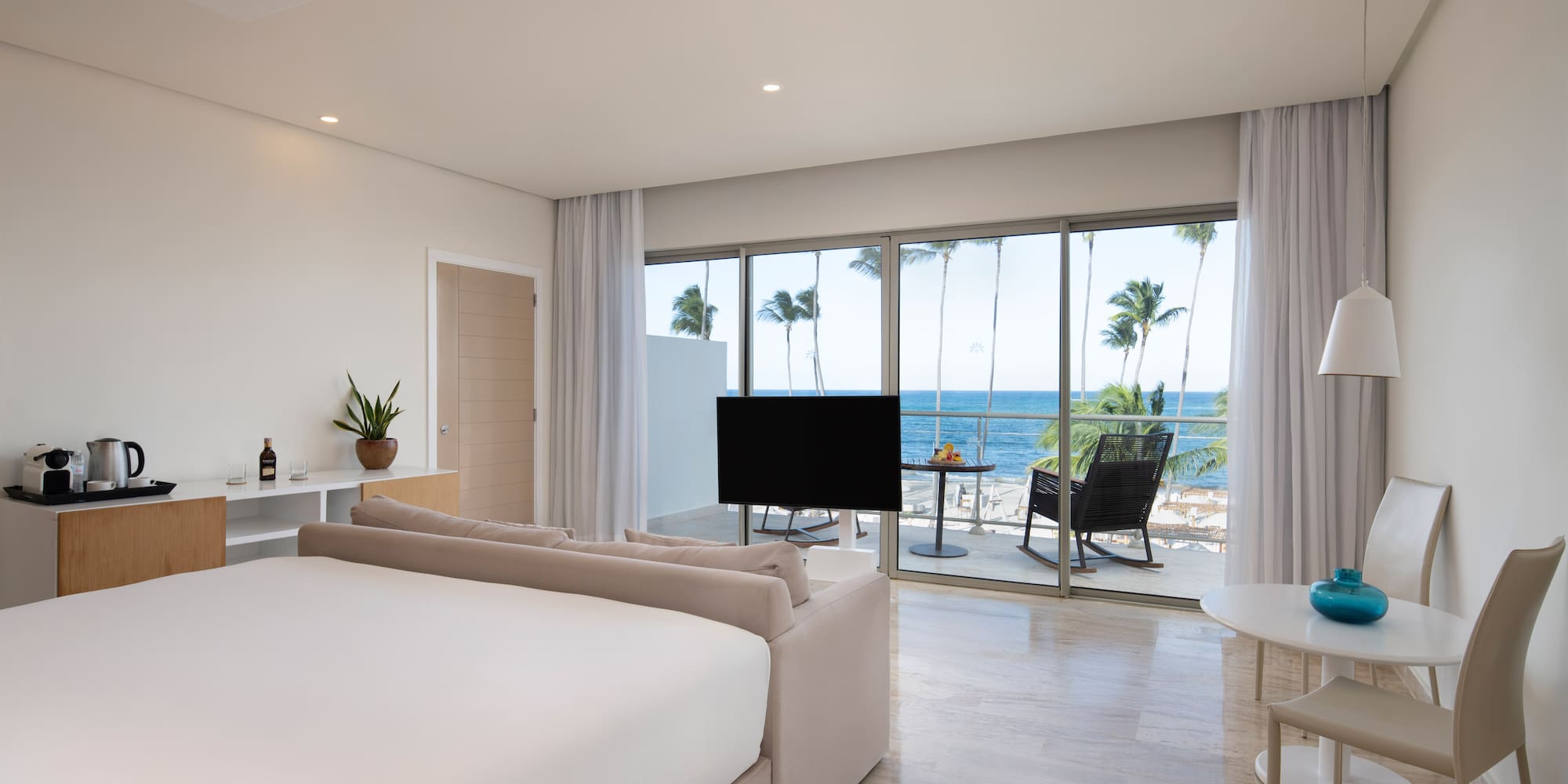 Modern hotel room with ocean view, balcony, palm trees, large bed, and lounge area.