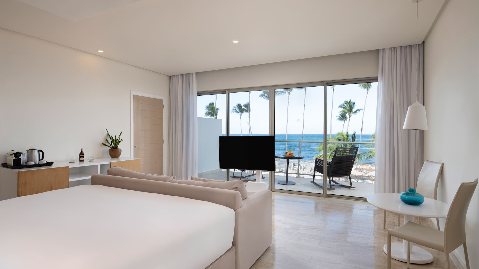 Modern hotel room with ocean view, balcony, palm trees, large bed, and lounge area.