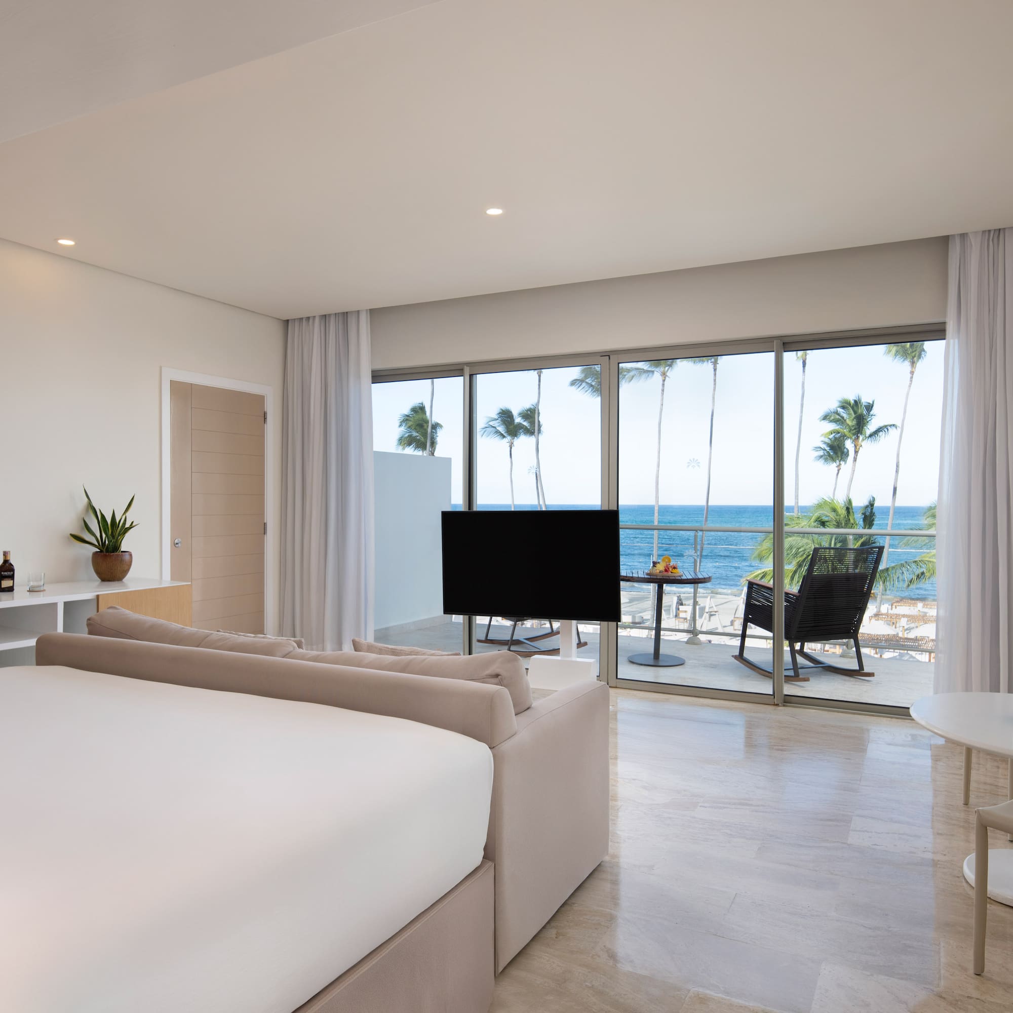Modern hotel room with ocean view, balcony, palm trees, large bed, and lounge area.