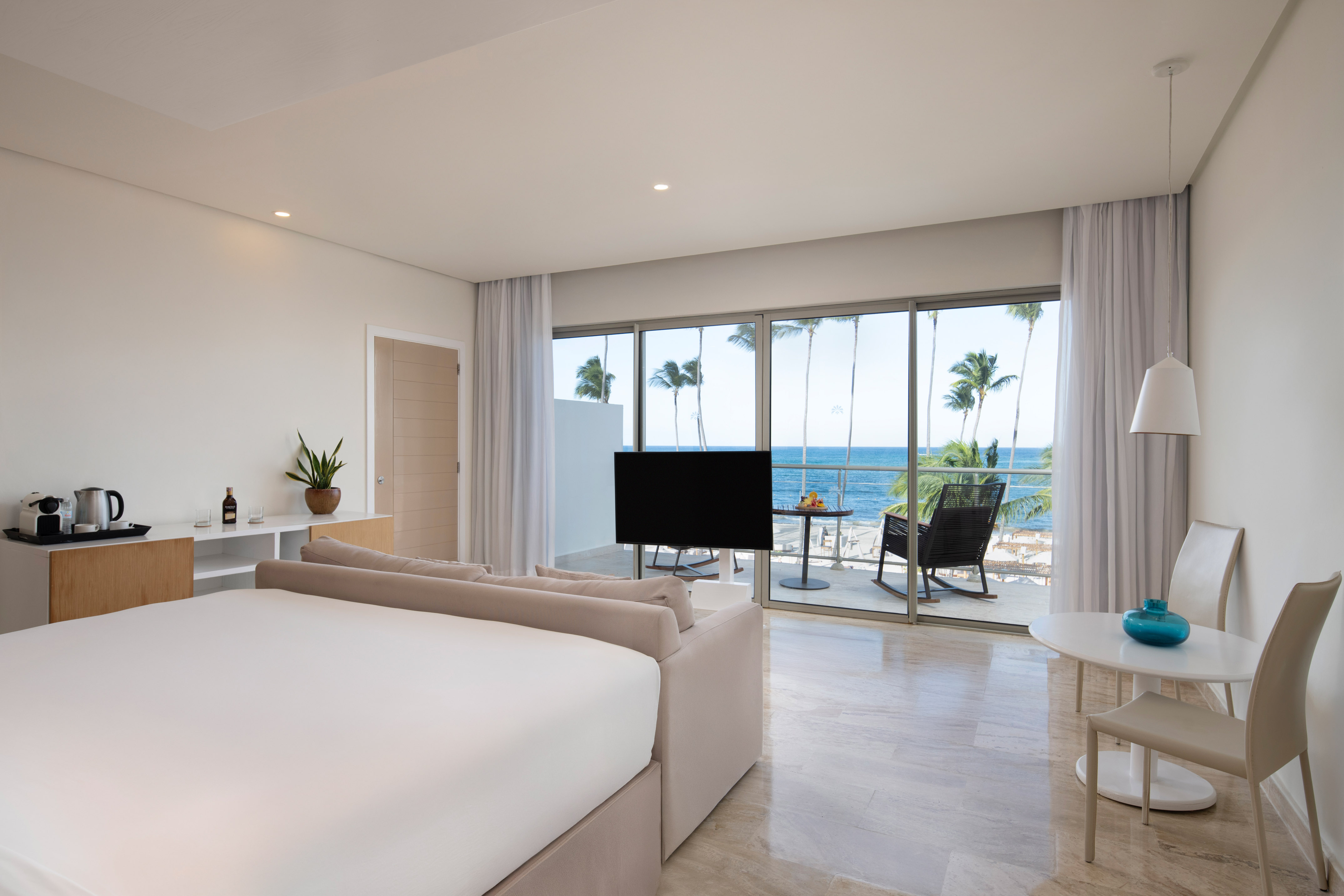 Modern hotel room with ocean view, balcony, palm trees, large bed, and lounge area.