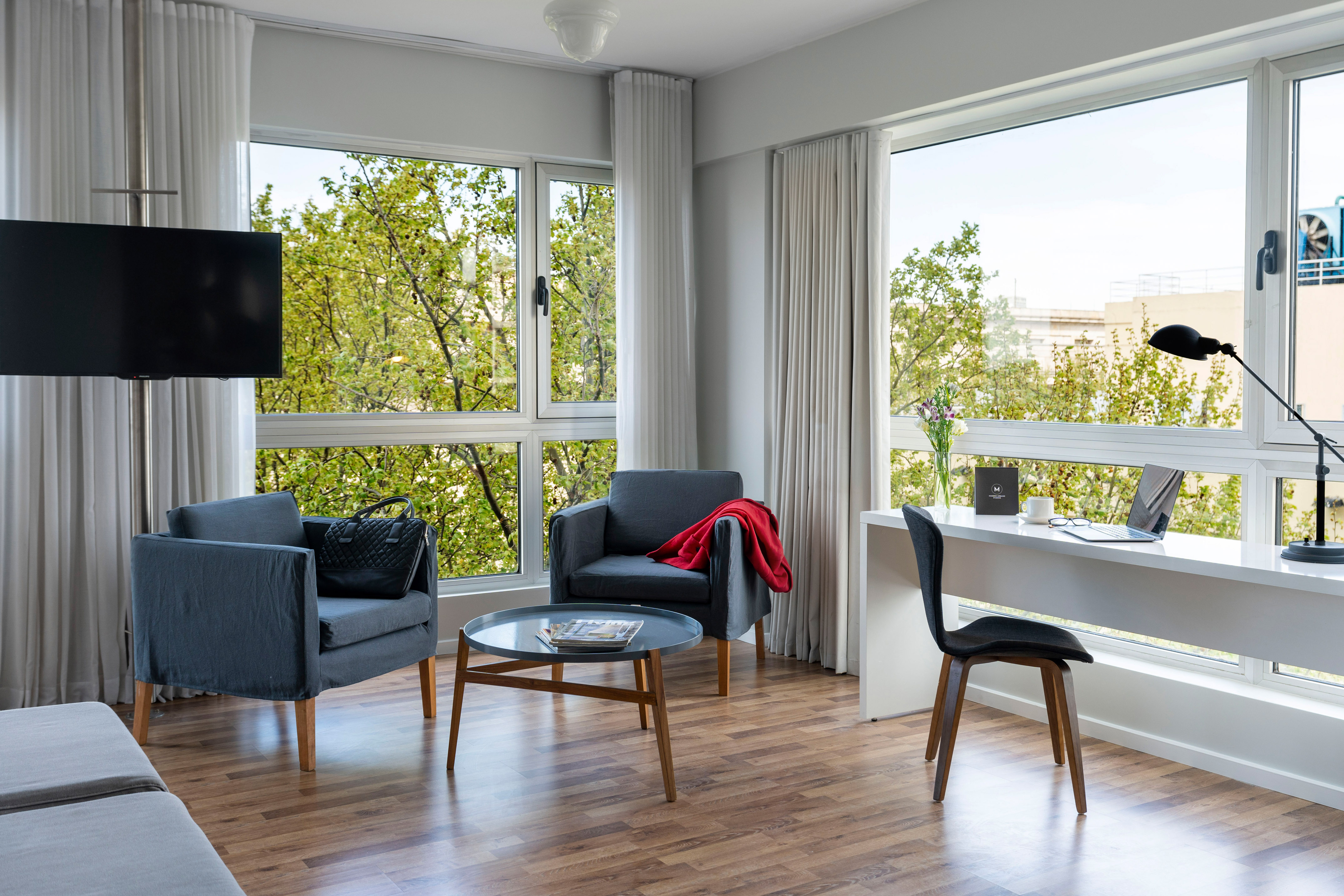 Bright modern living room with large windows, green trees, armchairs, desk, and laptop.
