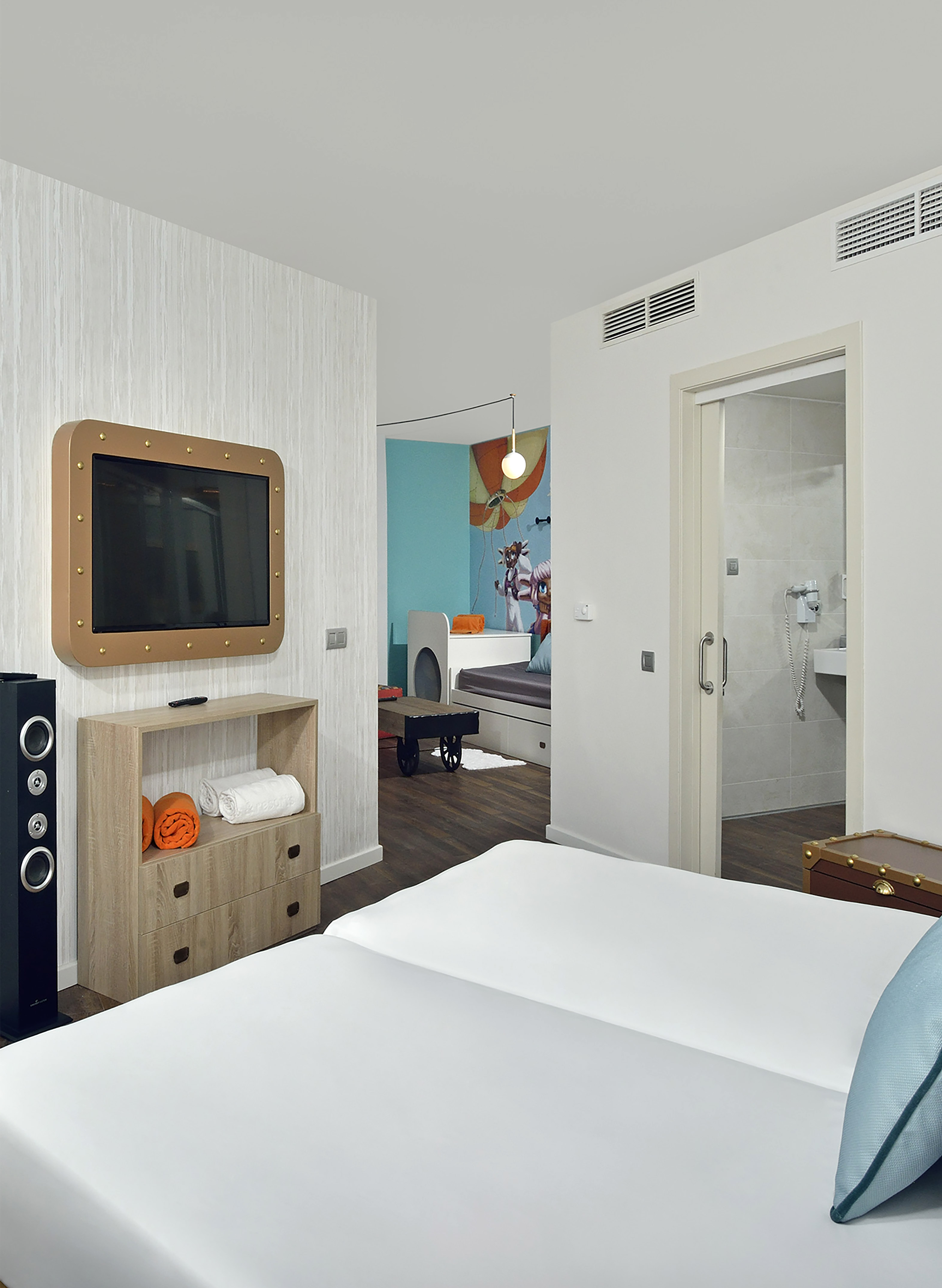 Hotel family room with white bed, TV, dresser, and colorful kids' area.