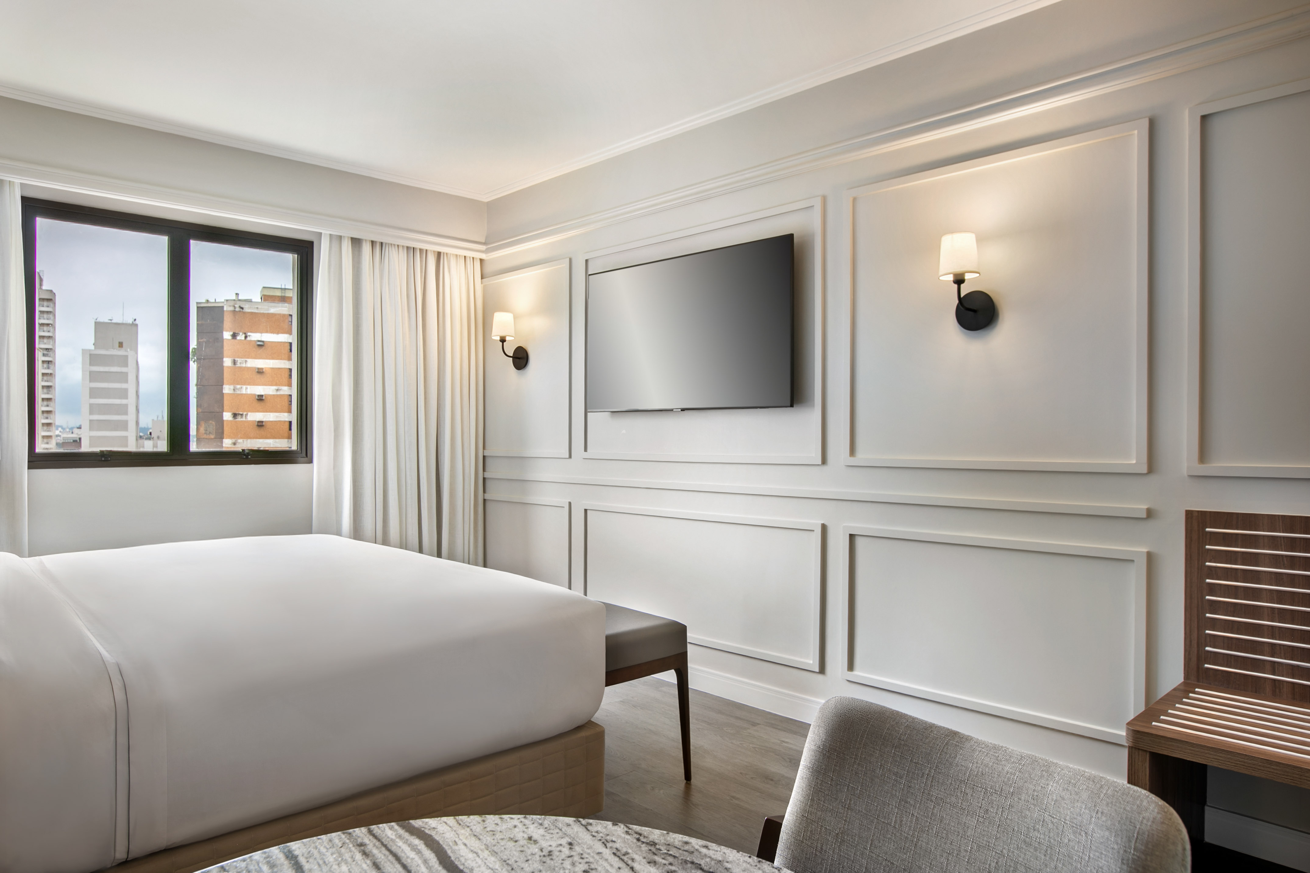 Modern hotel bedroom with white bed, city view, wall-mounted TV, and panel decor.