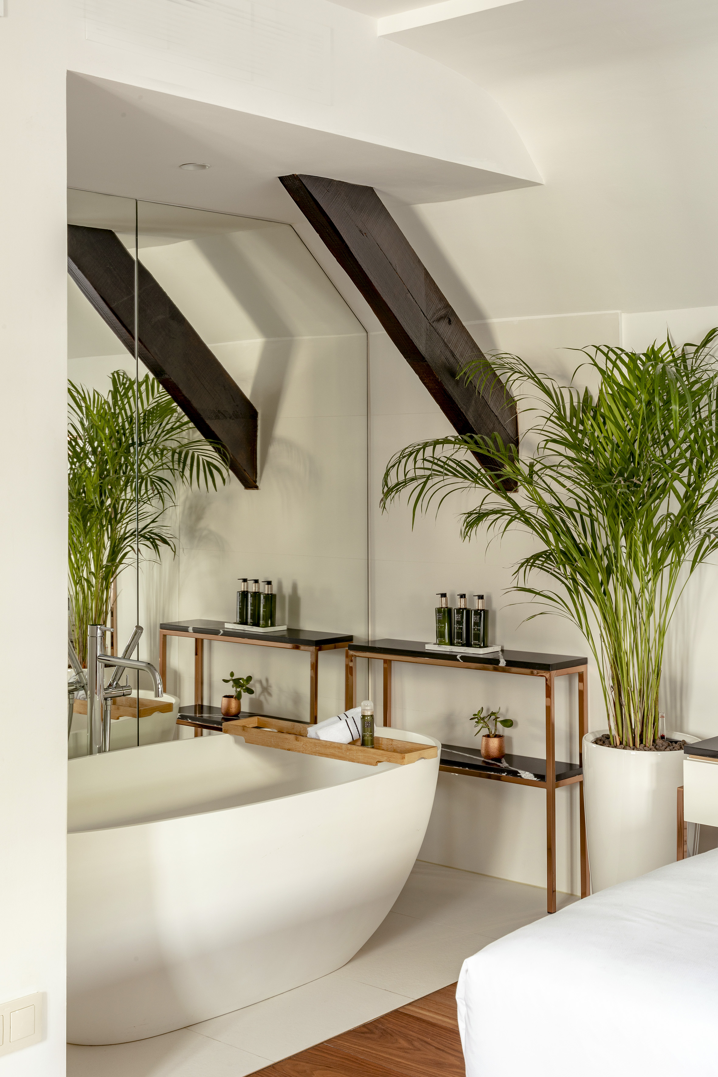 Modern bathroom suite with freestanding tub, mirror, large plant, and wood beams.