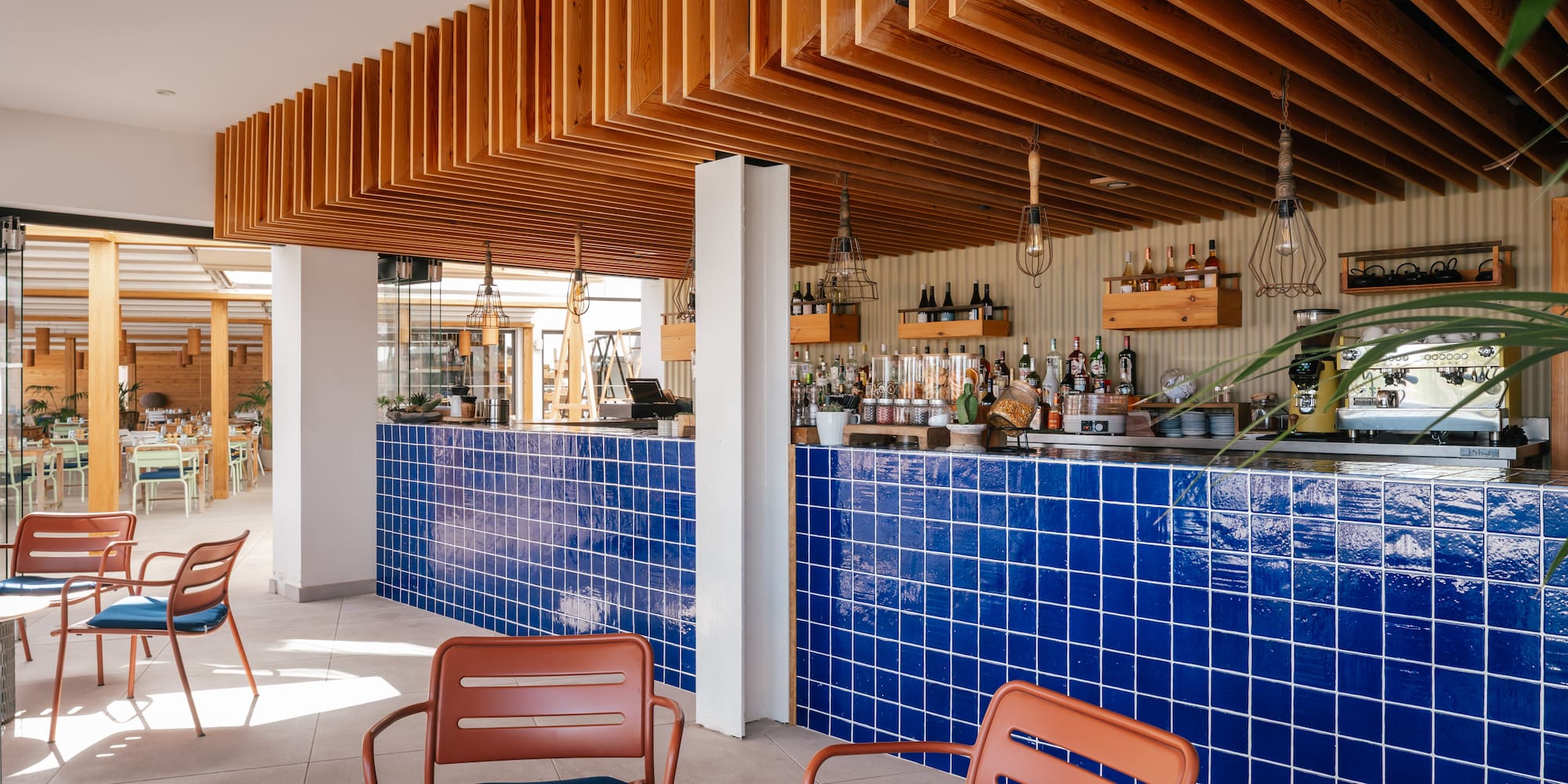 Modern bar with blue tiled counter, wood slat ceiling, and orange chairs.