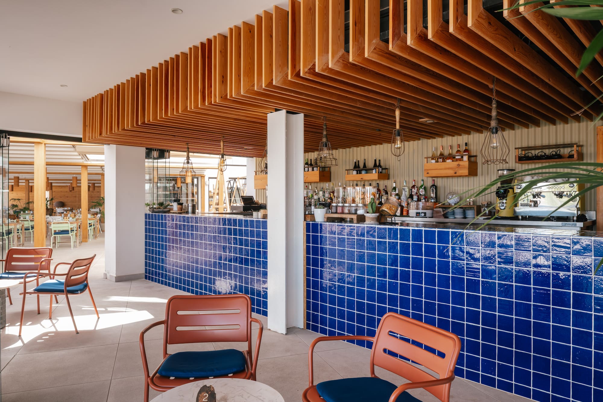 Modern bar with blue tiled counter, wood slat ceiling, and orange chairs.