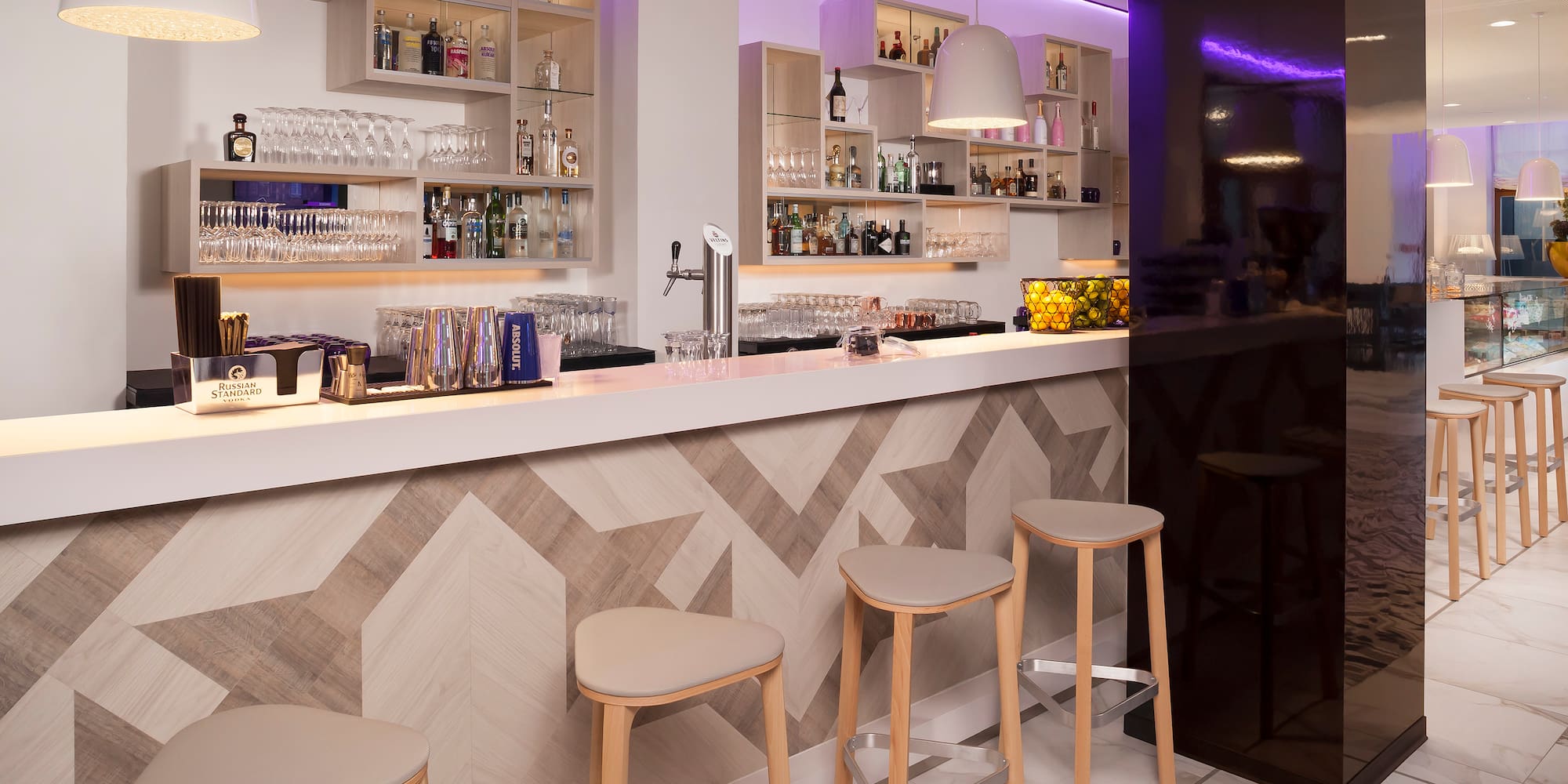 Modern hotel bar with chevron counter, three stools, liquor, and purple lighting.