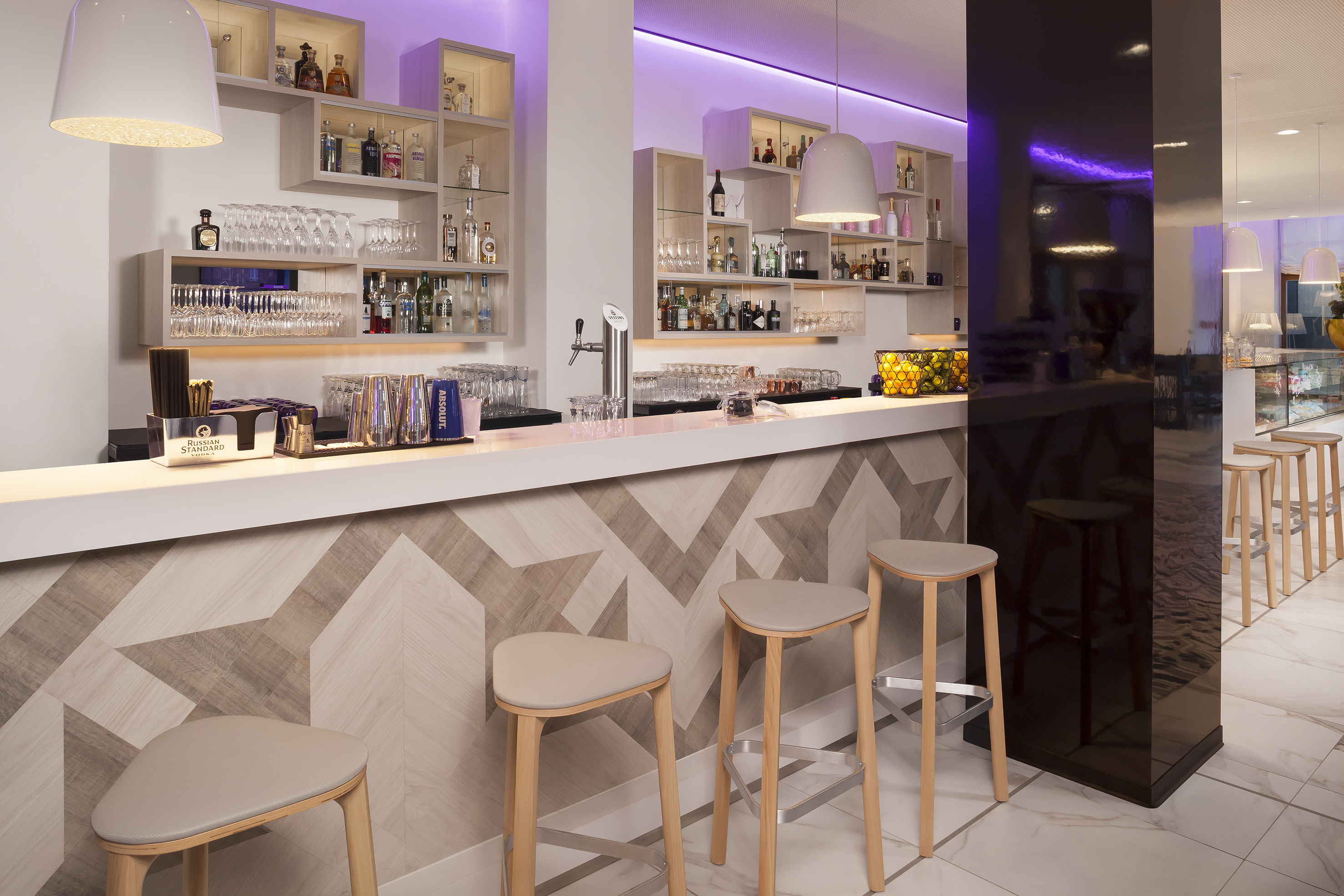 Modern hotel bar with chevron counter, three stools, liquor, and purple lighting.