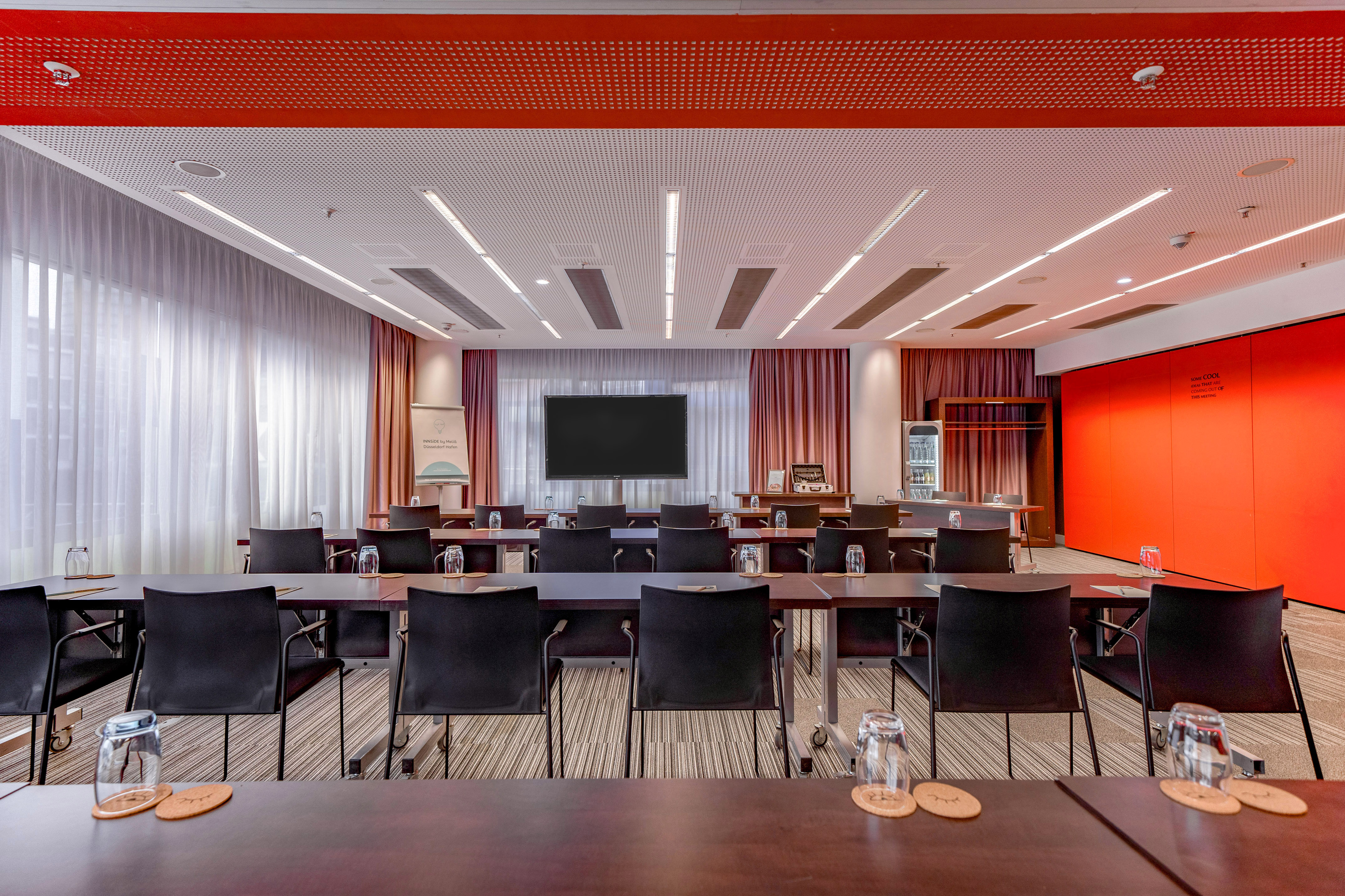 Modern meeting room with tables, black chairs, large screen, red ceiling.