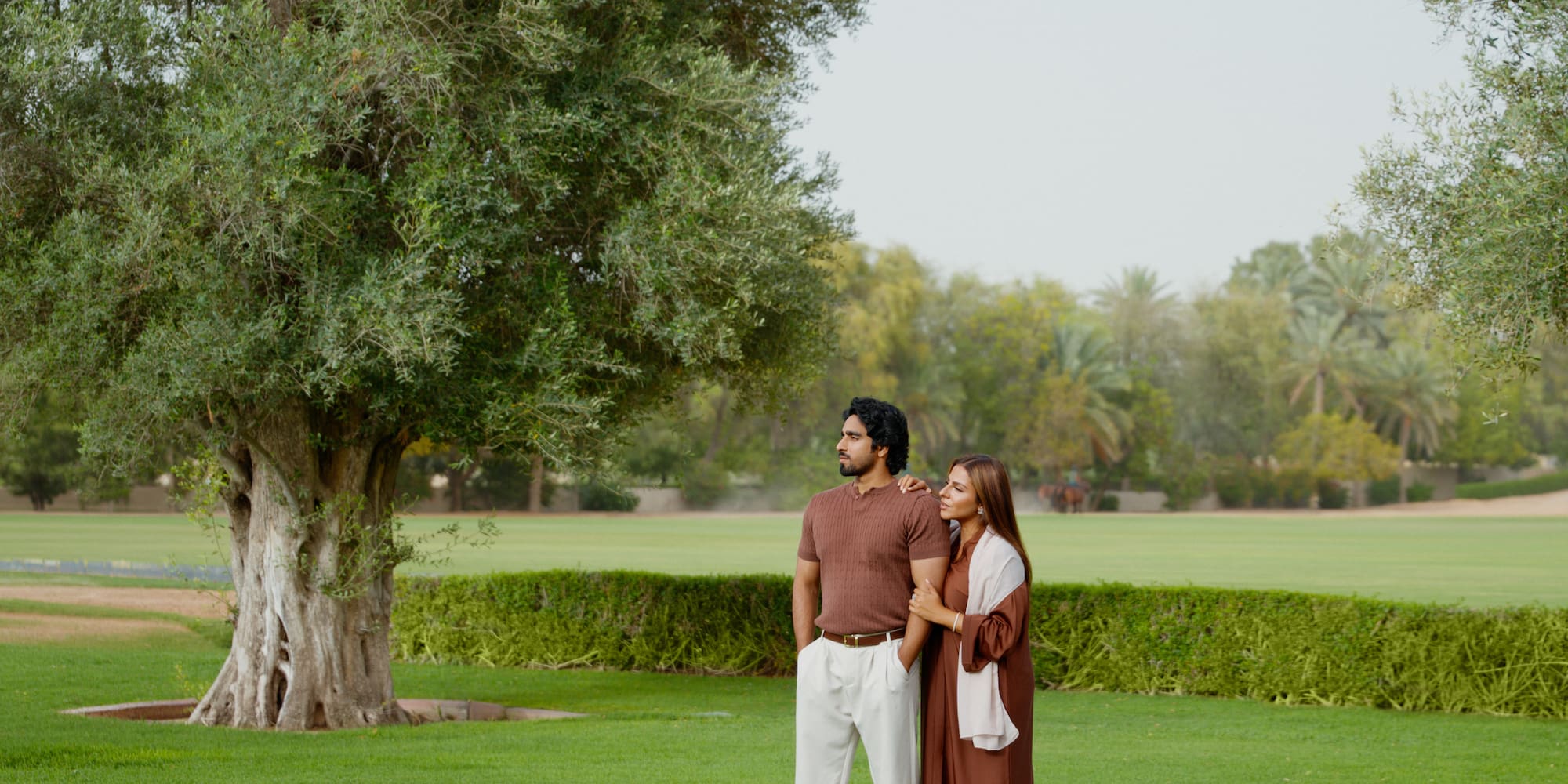 Couple in brown abaya and polo shirt standing in a lush green park.