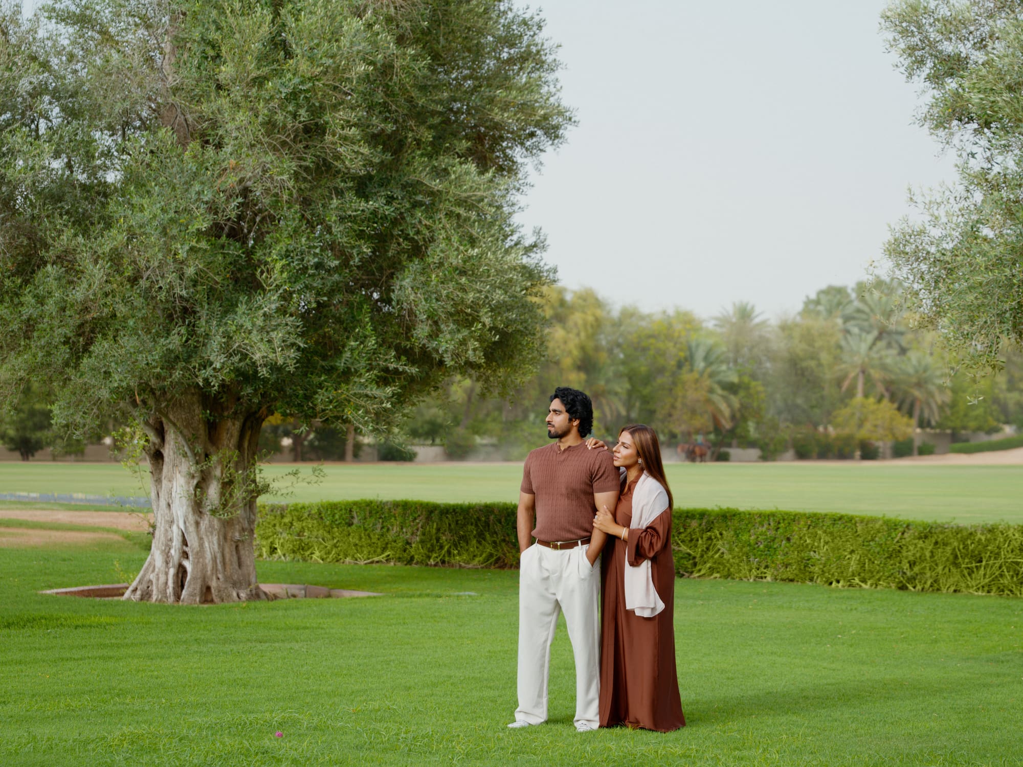 Couple in brown abaya and polo shirt standing in a lush green park.