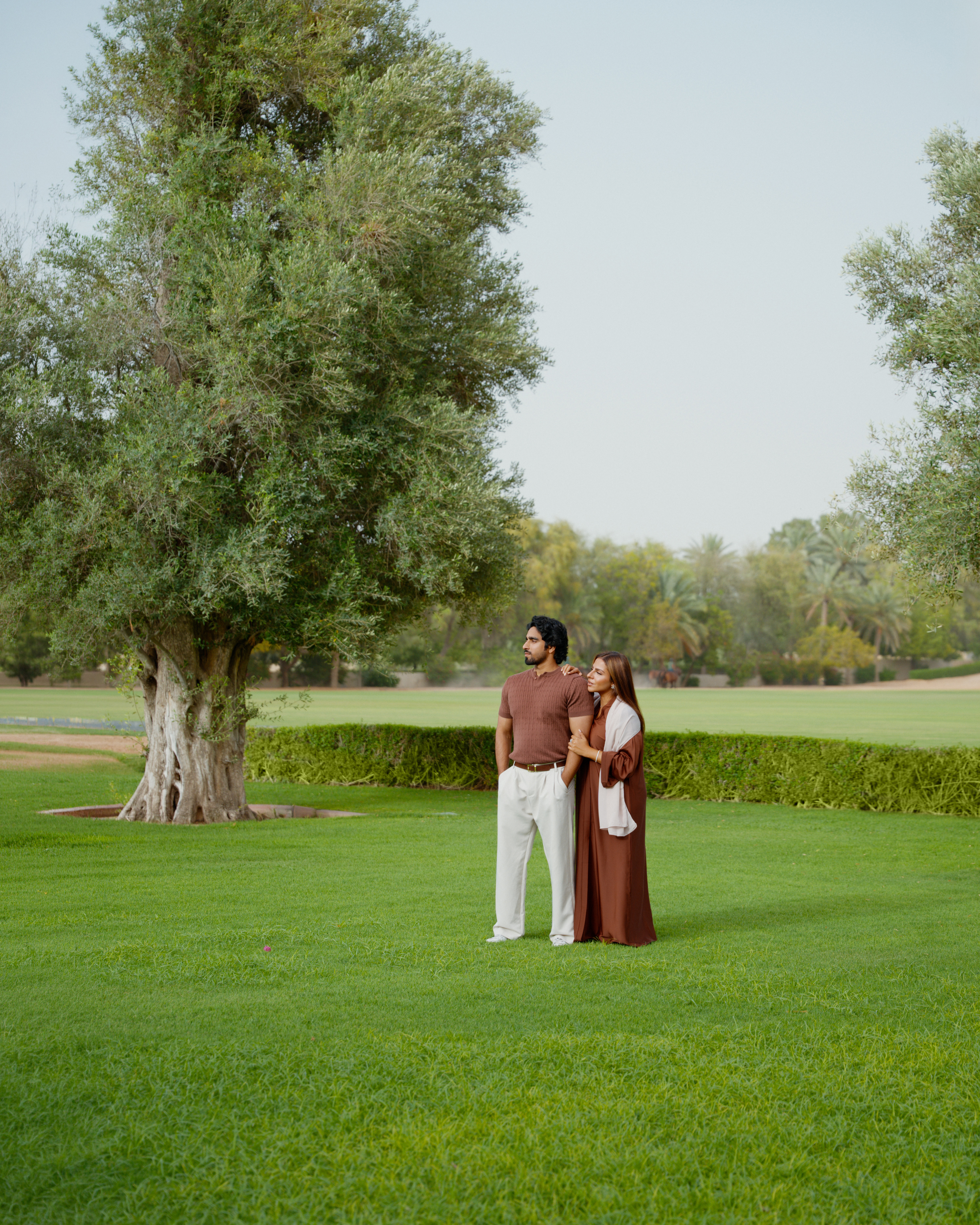 Couple in brown abaya and polo shirt standing in a lush green park.