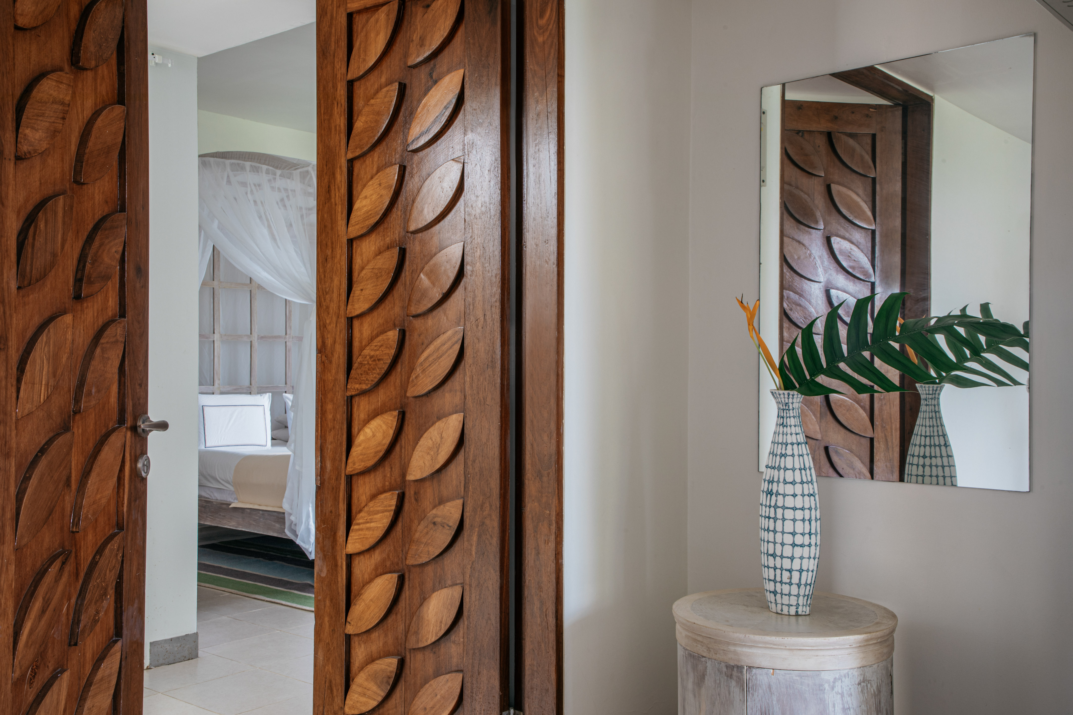 Wooden leaf-carved door leading to tropical bedroom with canopy bed and vase.