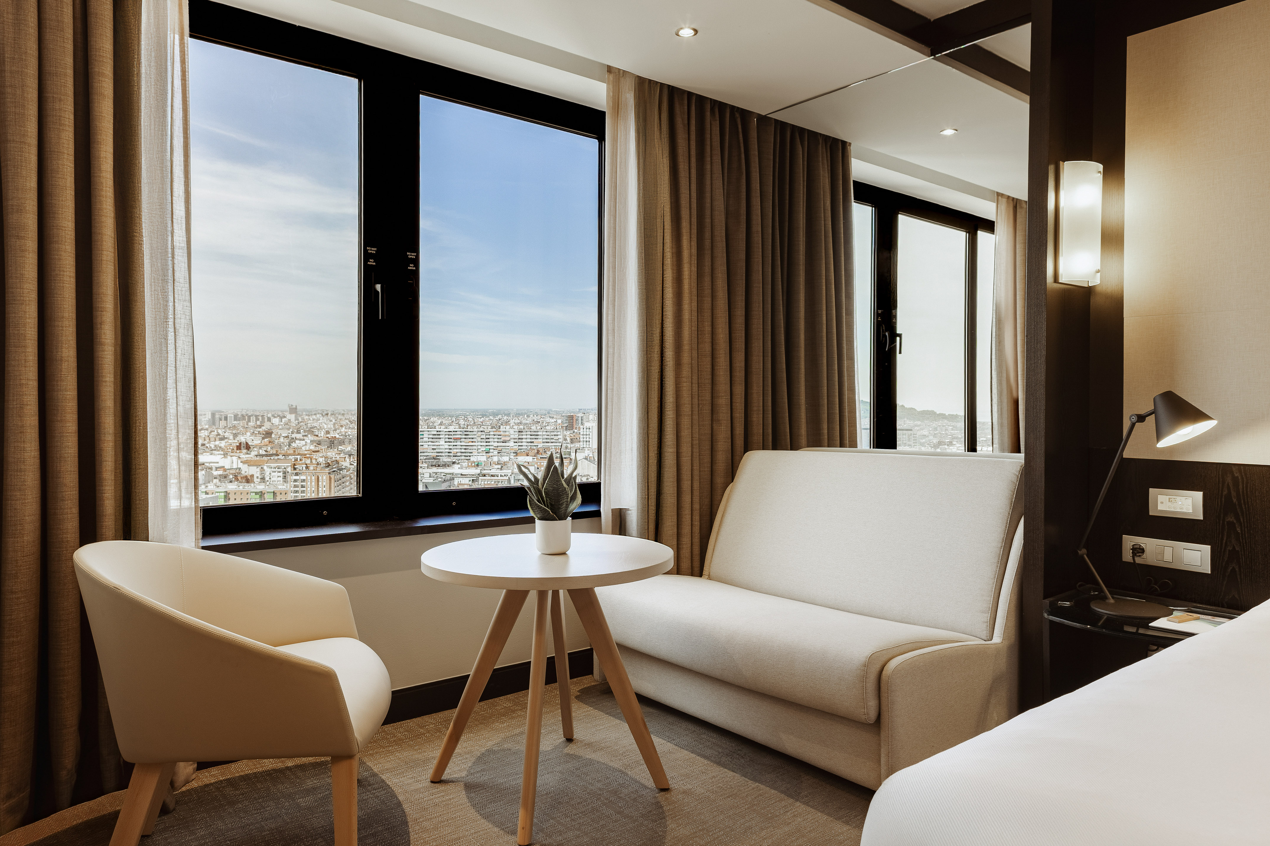 Modern hotel suite with city view, large windows, beige sofa and armchair.