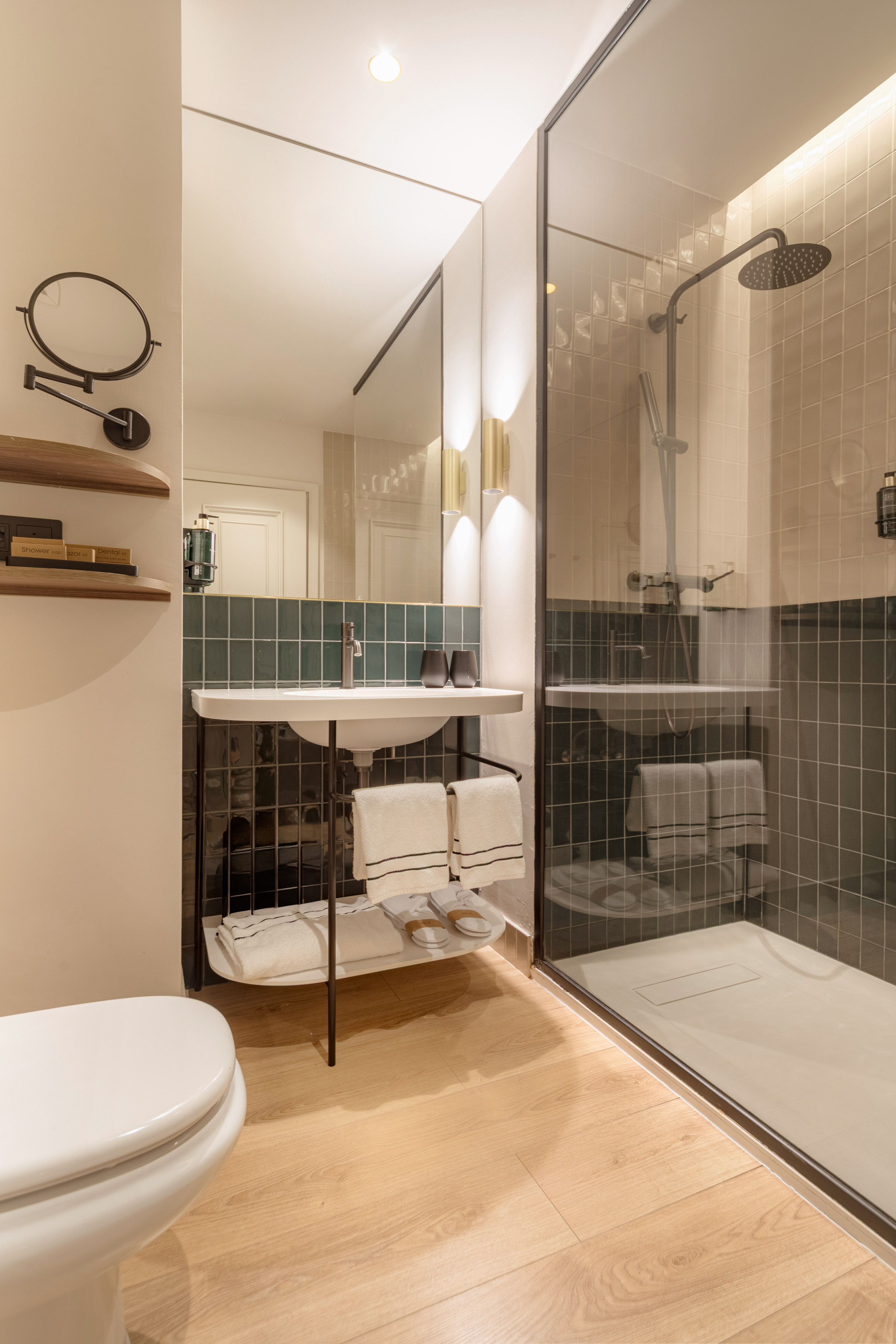 Modern bathroom with toilet, white sink, dark green tiled wall, glass shower, wood floor.
