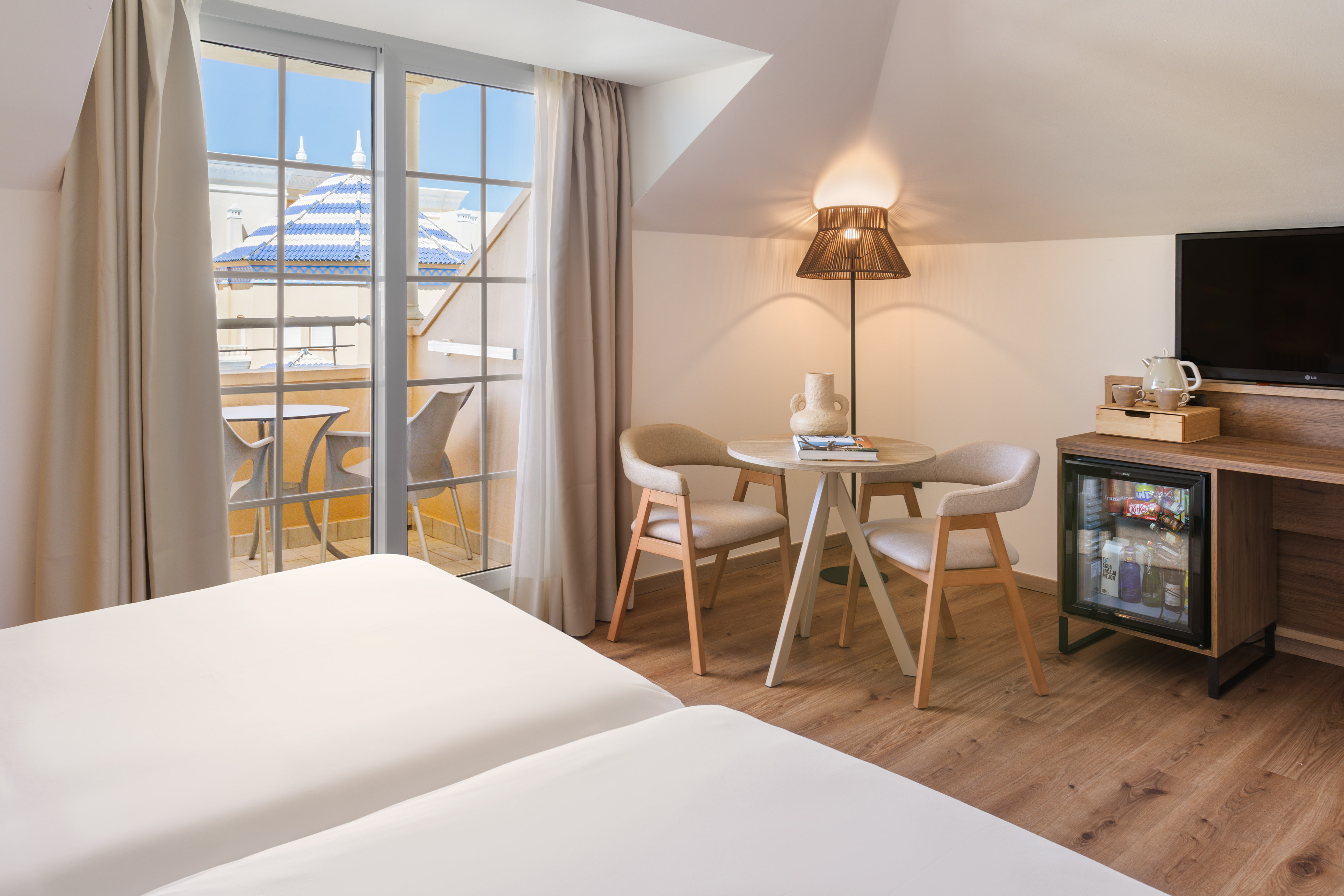 Bright hotel room with twin beds, balcony, table, chairs, and minibar.
