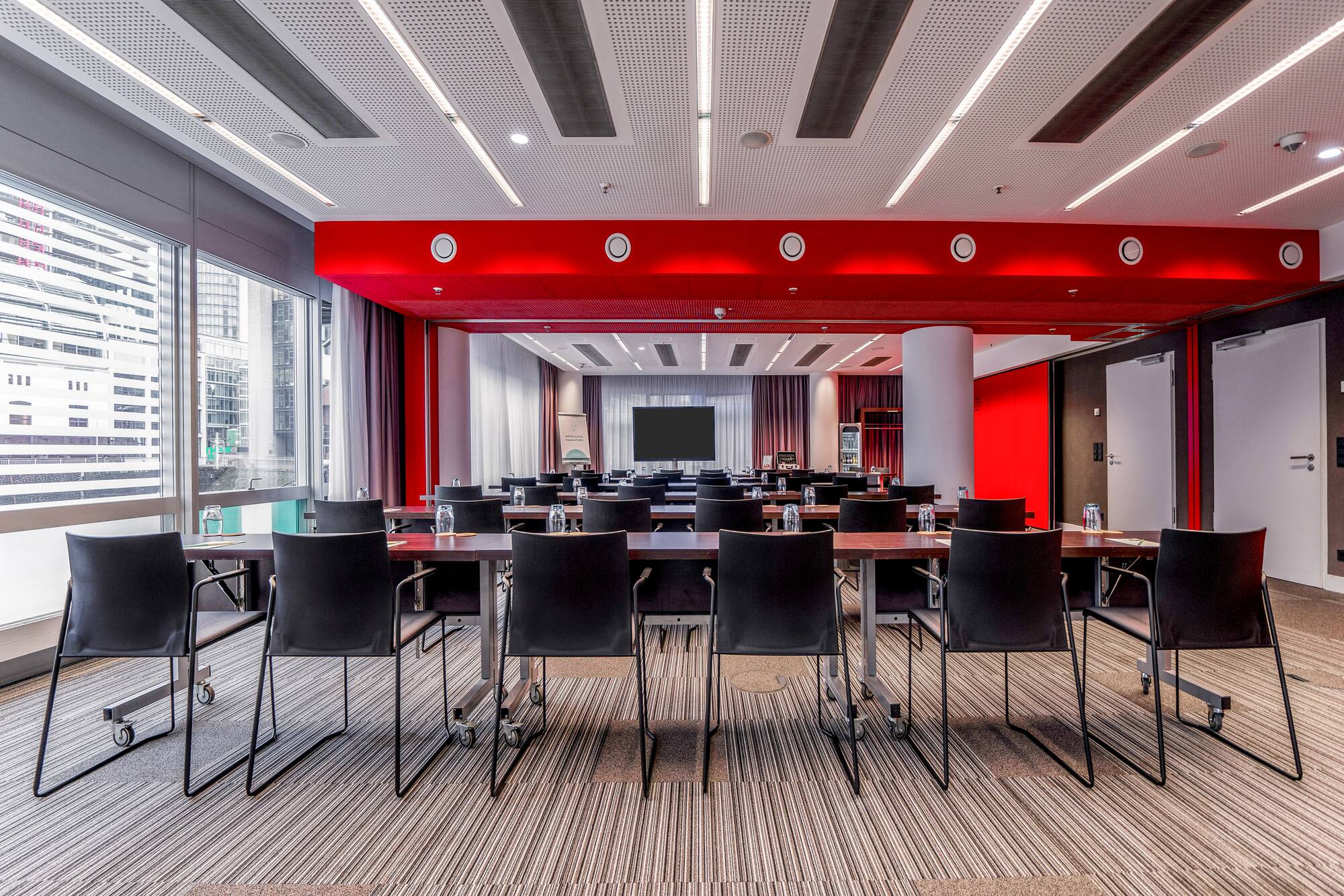 Modern meeting room with red ceiling, city windows, tables, and large screen.