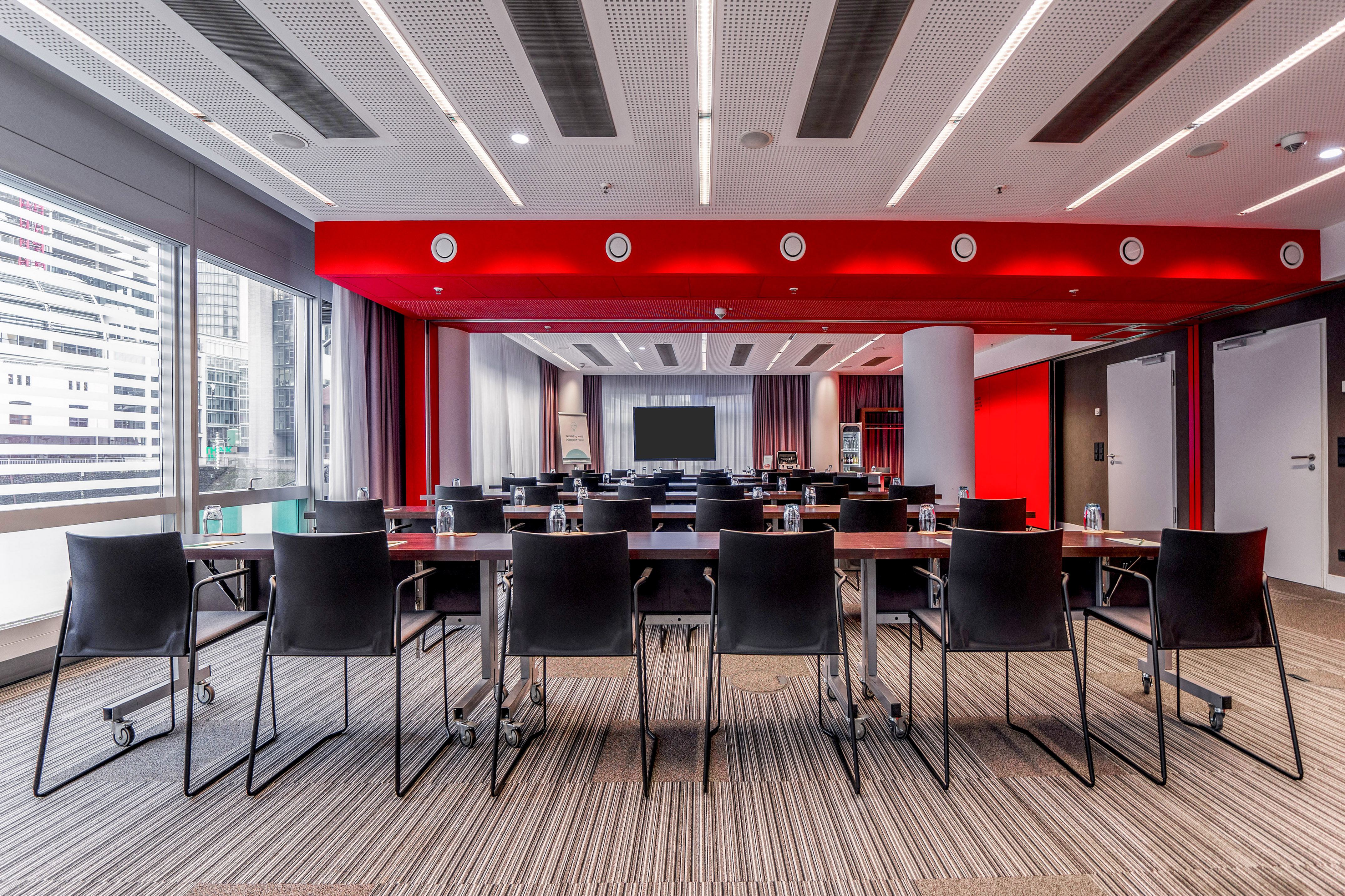 Modern meeting room with red ceiling, city windows, tables, and large screen.