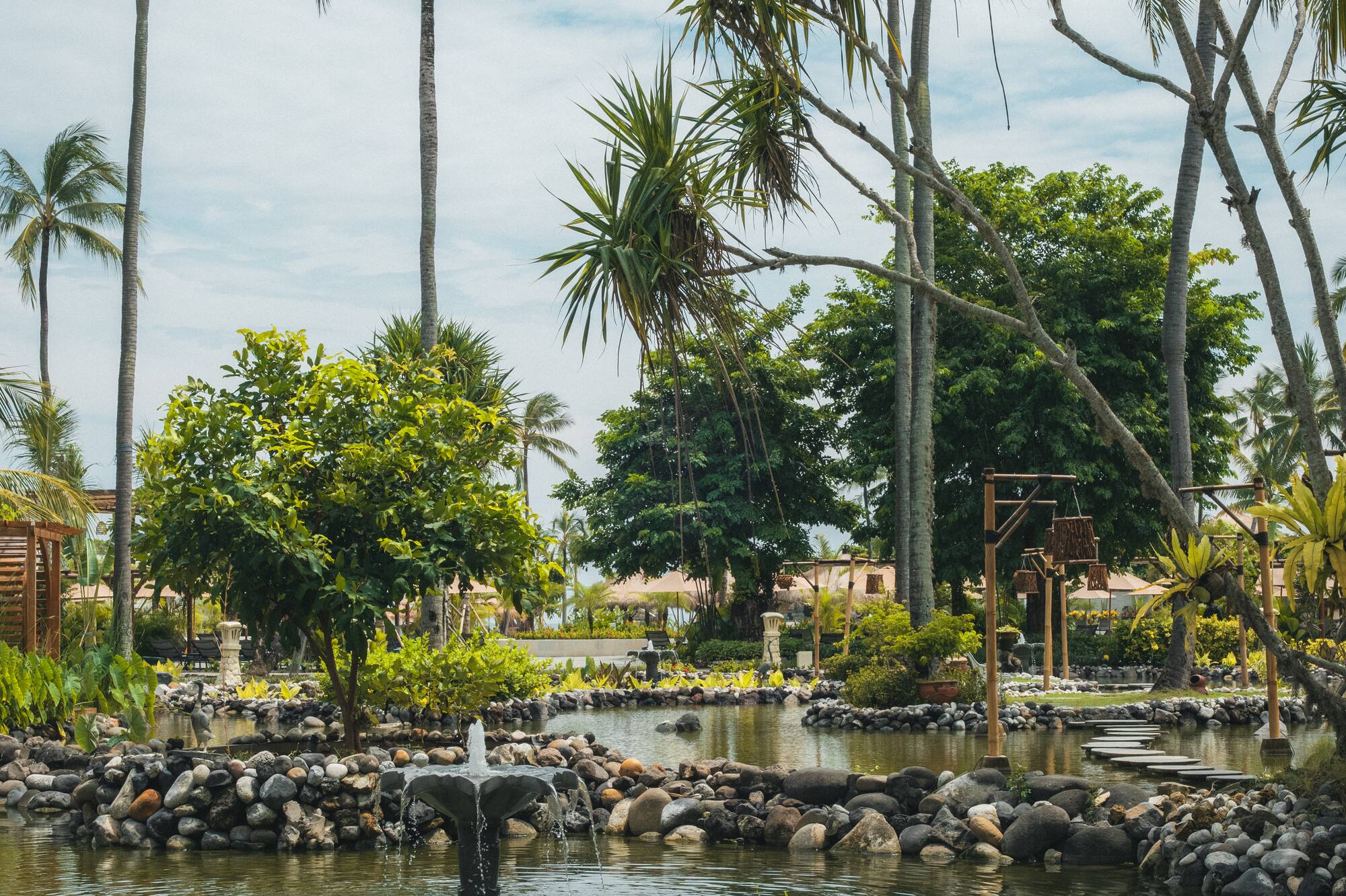 Estanque tropical con fuente, palmeras y exuberante vegetación bajo cielo claro.