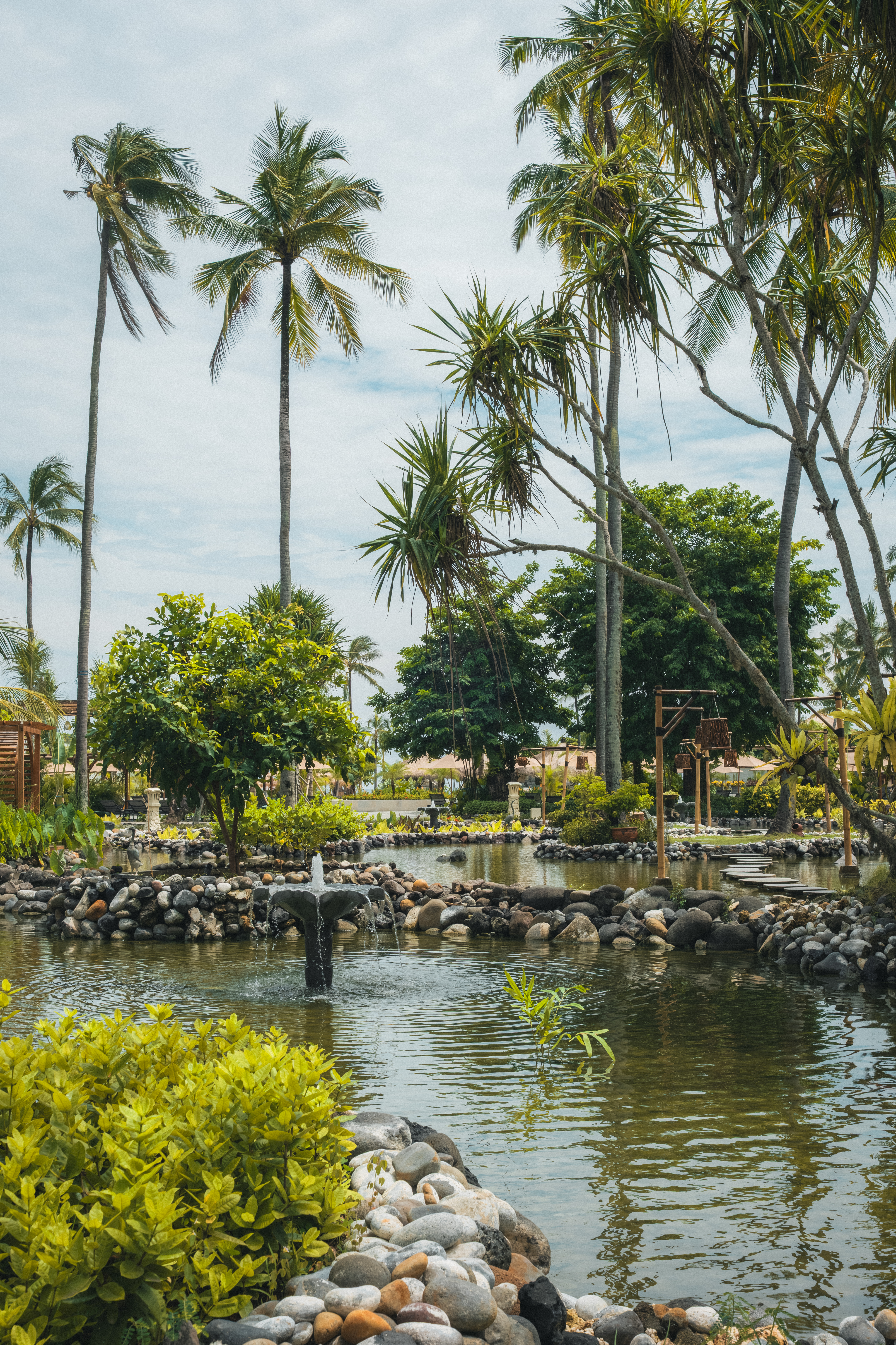 Estanque tropical con fuente, palmeras y exuberante vegetación bajo cielo claro.