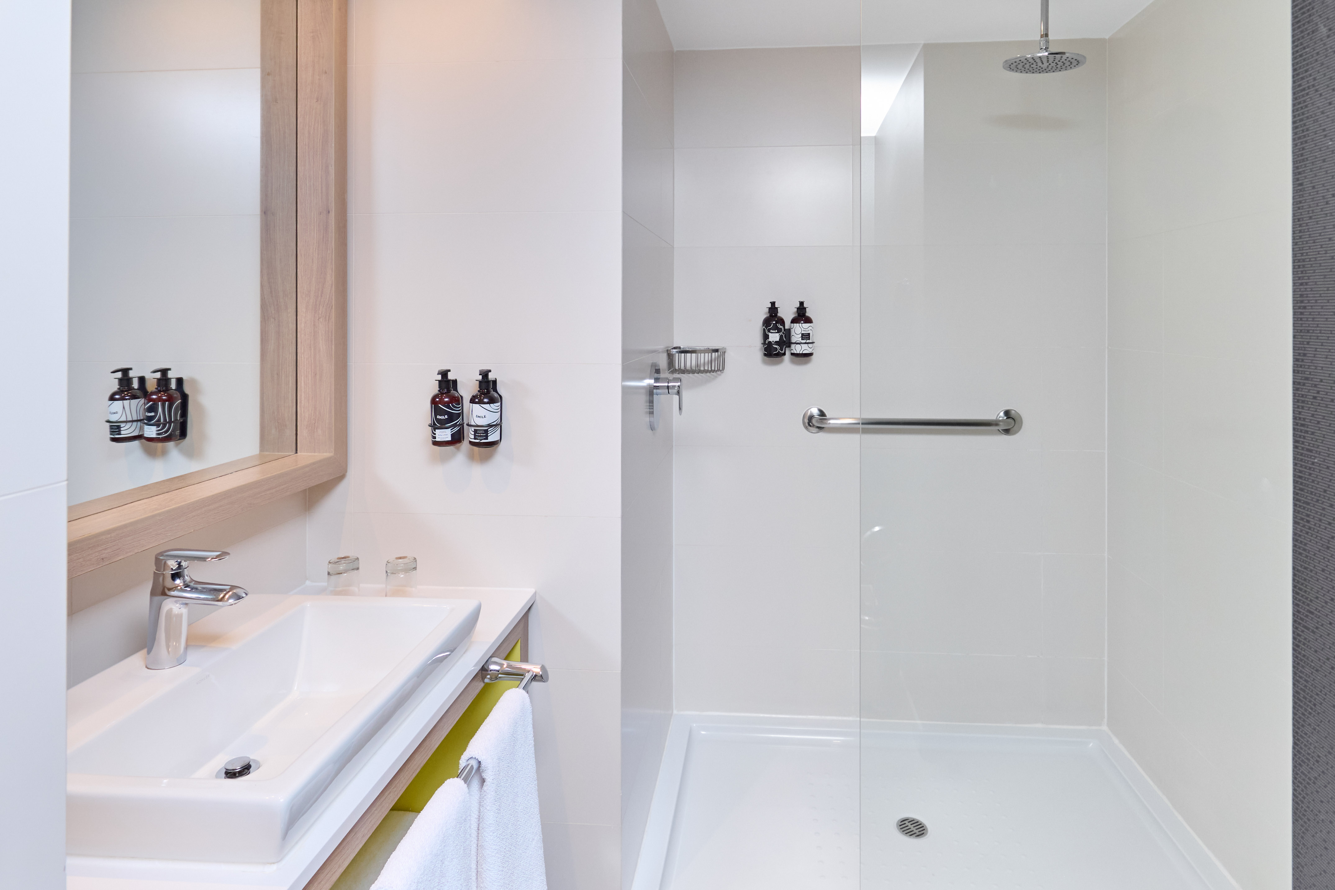 Modern hotel bathroom with white tiles, vanity sink, and walk-in shower with grab bar.