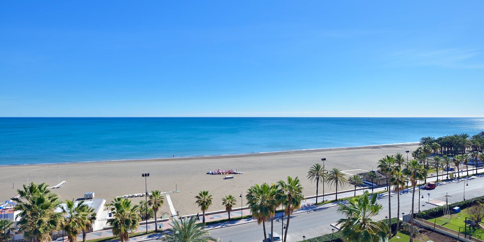 Sunny beach with blue ocean, palm trees, and coastal road.
