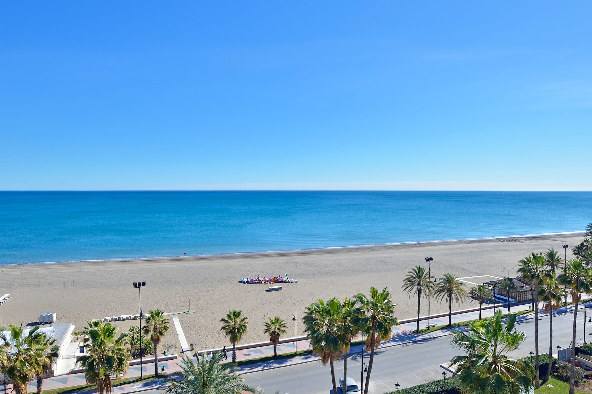 Sunny beach with blue ocean, palm trees, and coastal road.