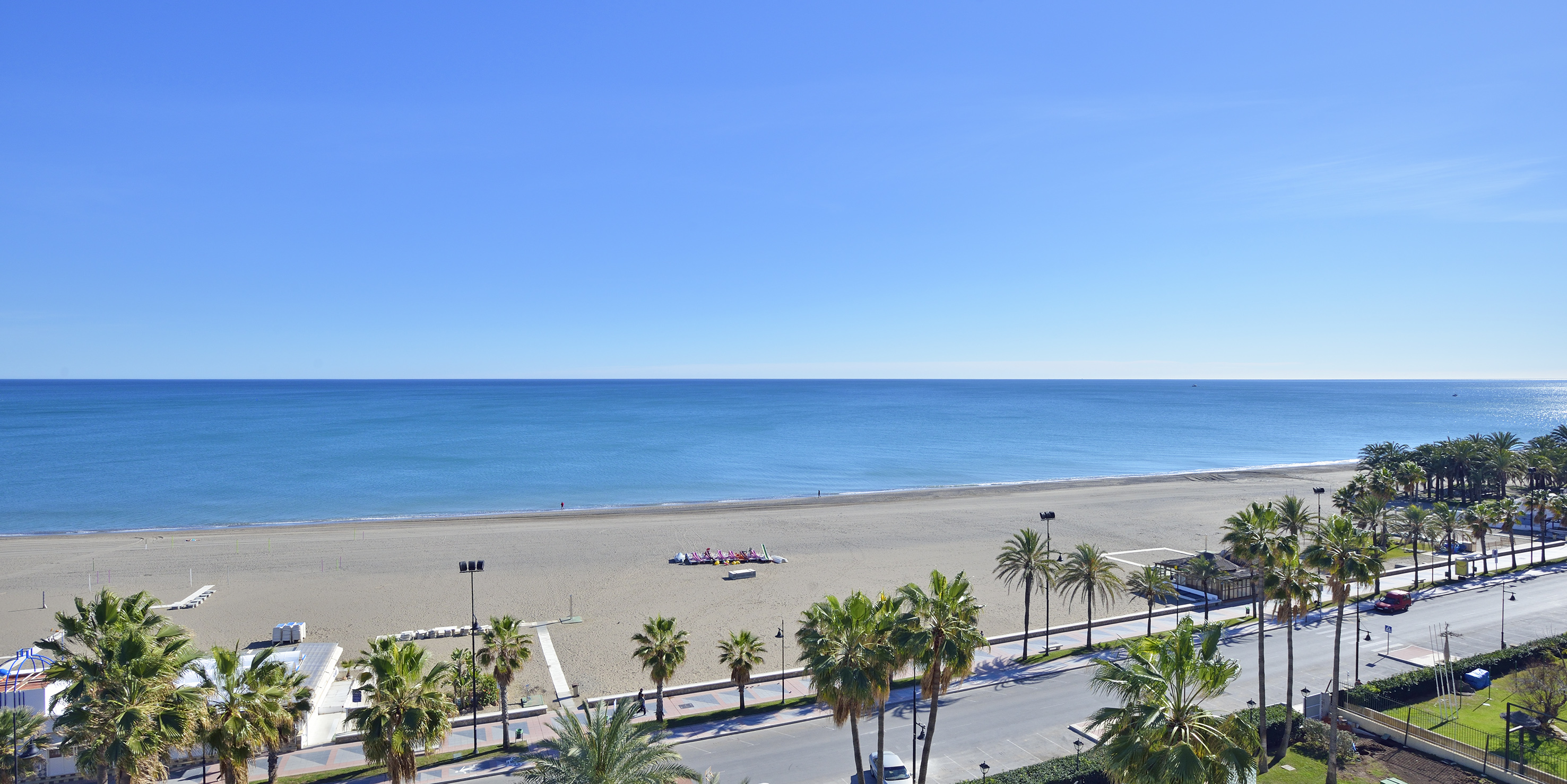 Sunny beach with blue ocean, palm trees, and coastal road.