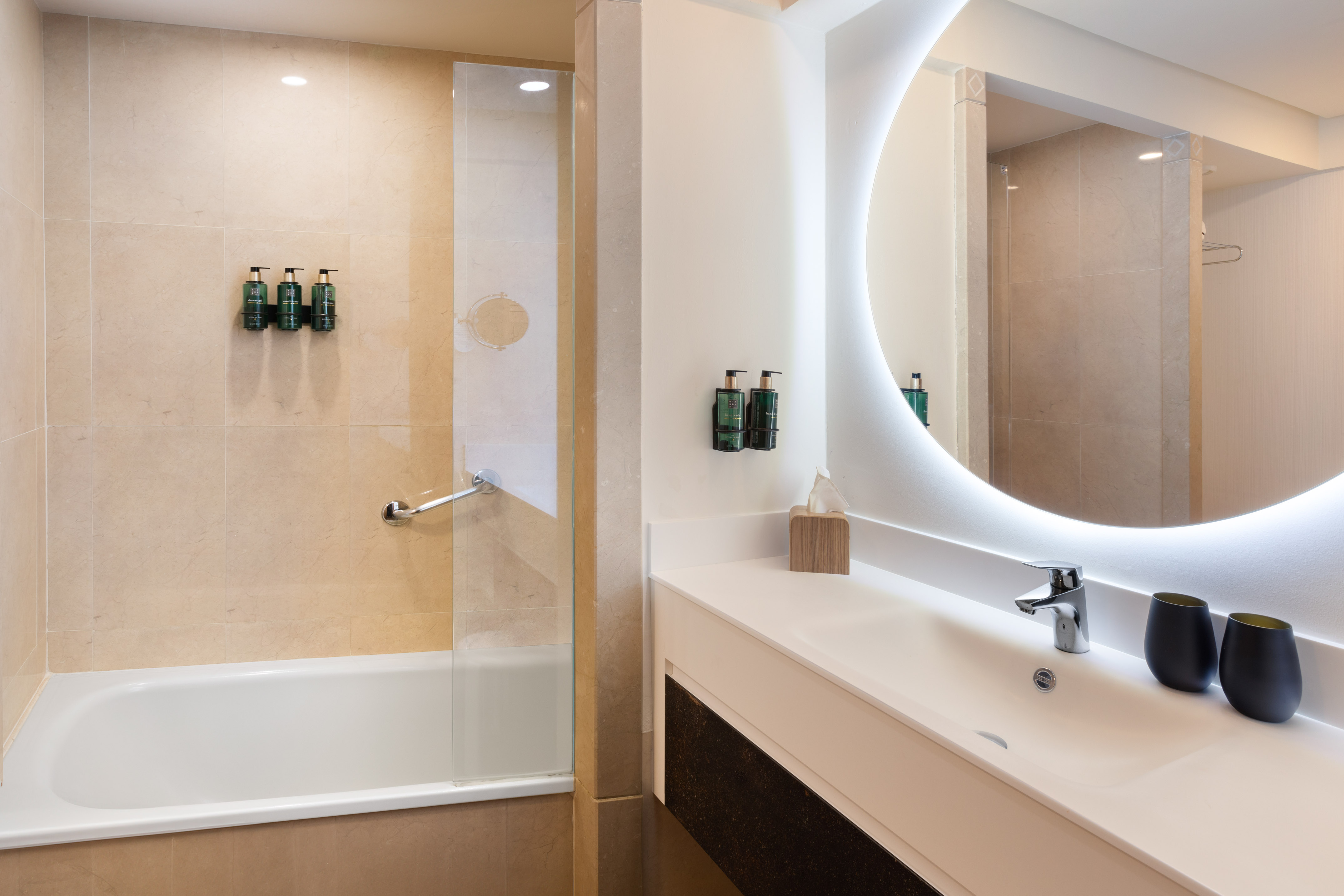 Hotel bathroom with bathtub, vanity sink, and backlit round mirror.