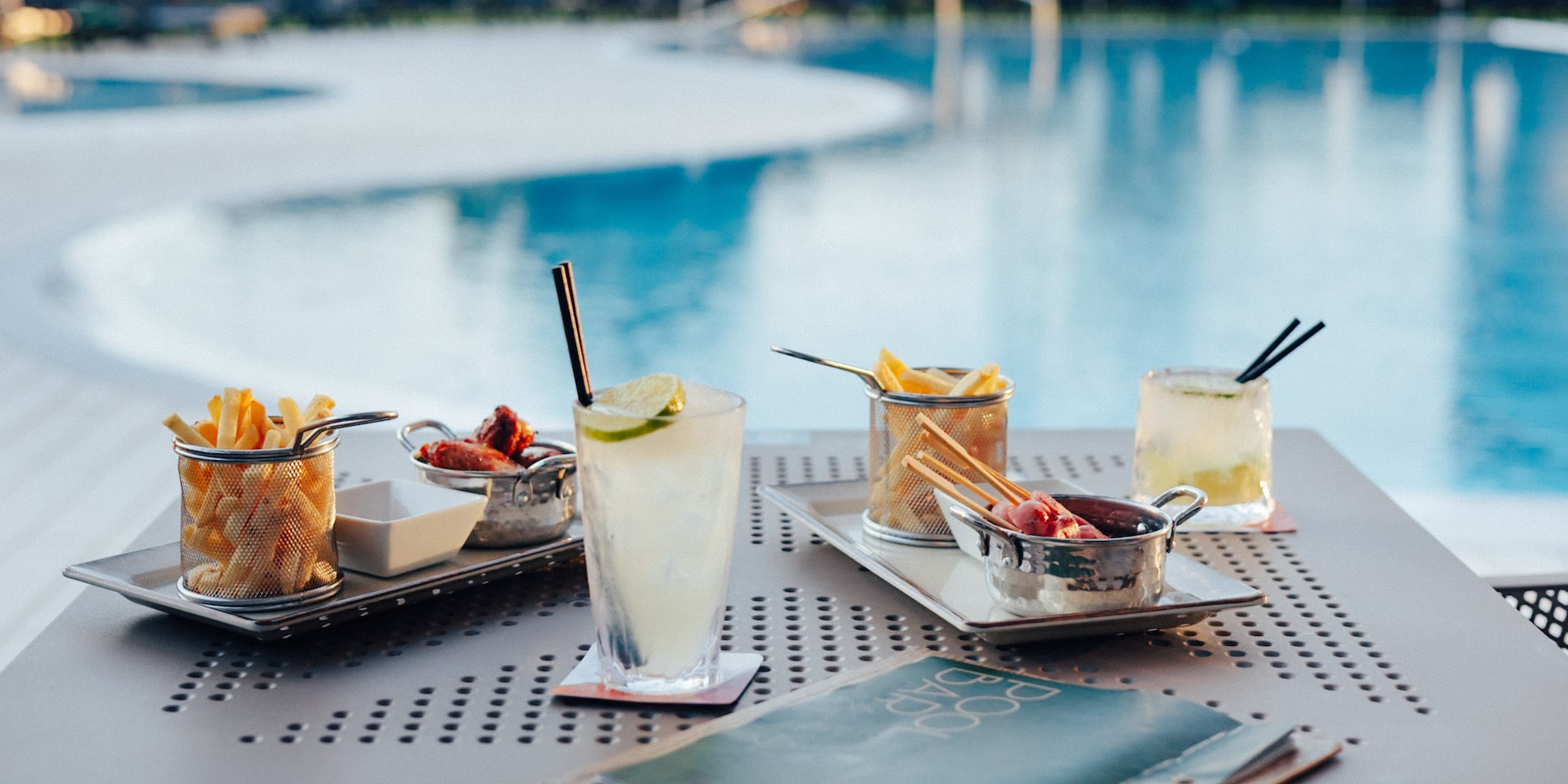 Cocktails, french fries, and chicken wings on a table by a resort pool.