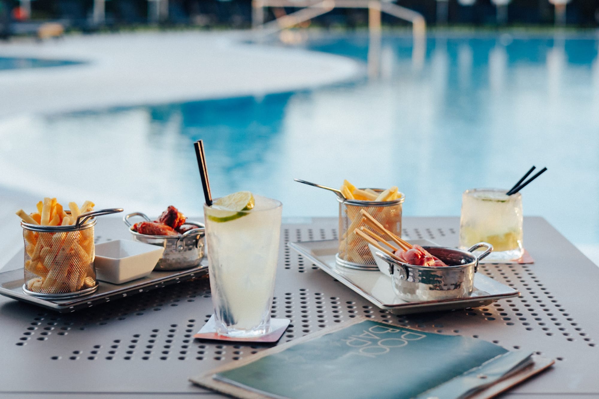 Cocktails, french fries, and chicken wings on a table by a resort pool.