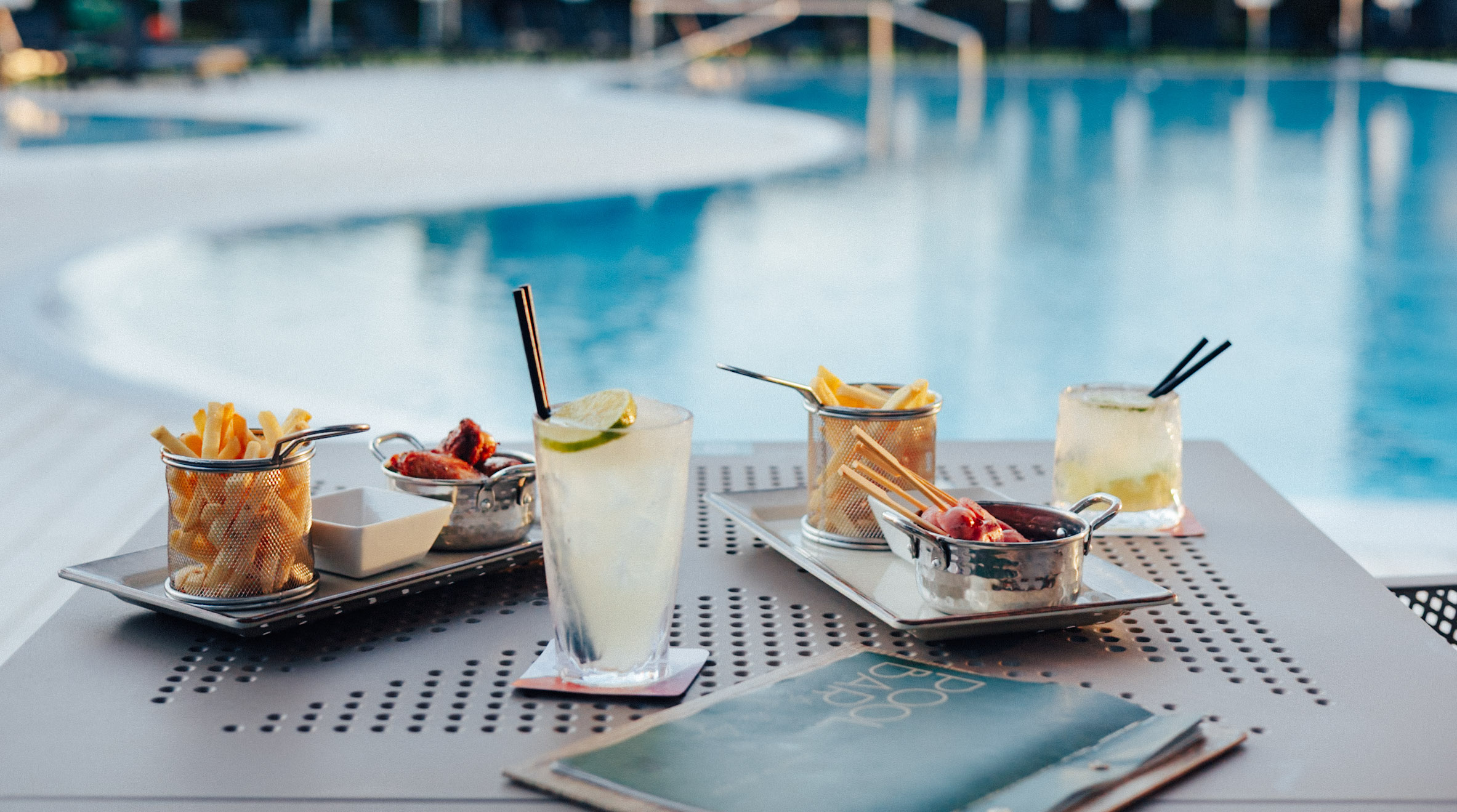 Cocktails, french fries, and chicken wings on a table by a resort pool.