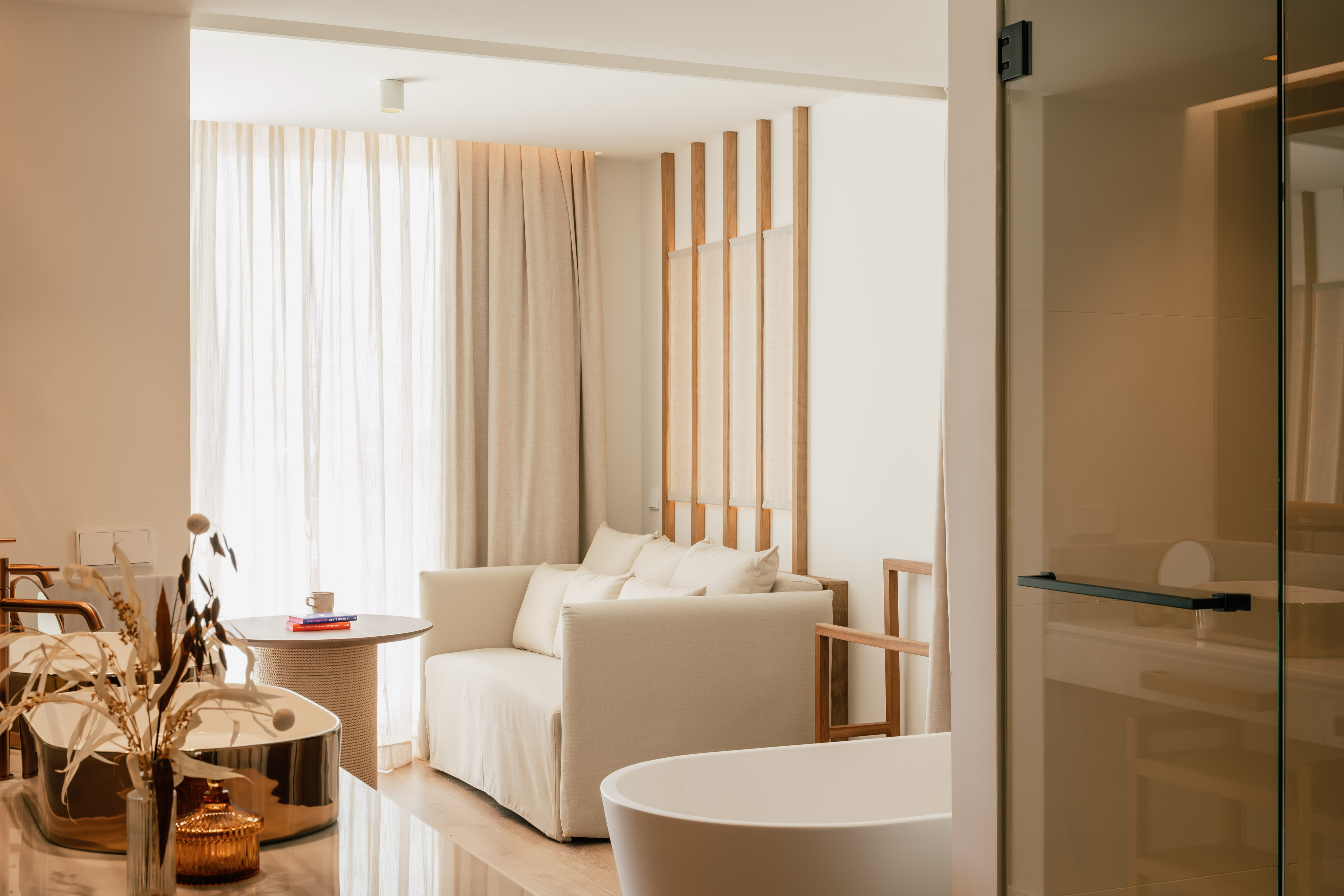 Modern luxury hotel suite with neutral sofa, sheer curtains, and freestanding bathtub.