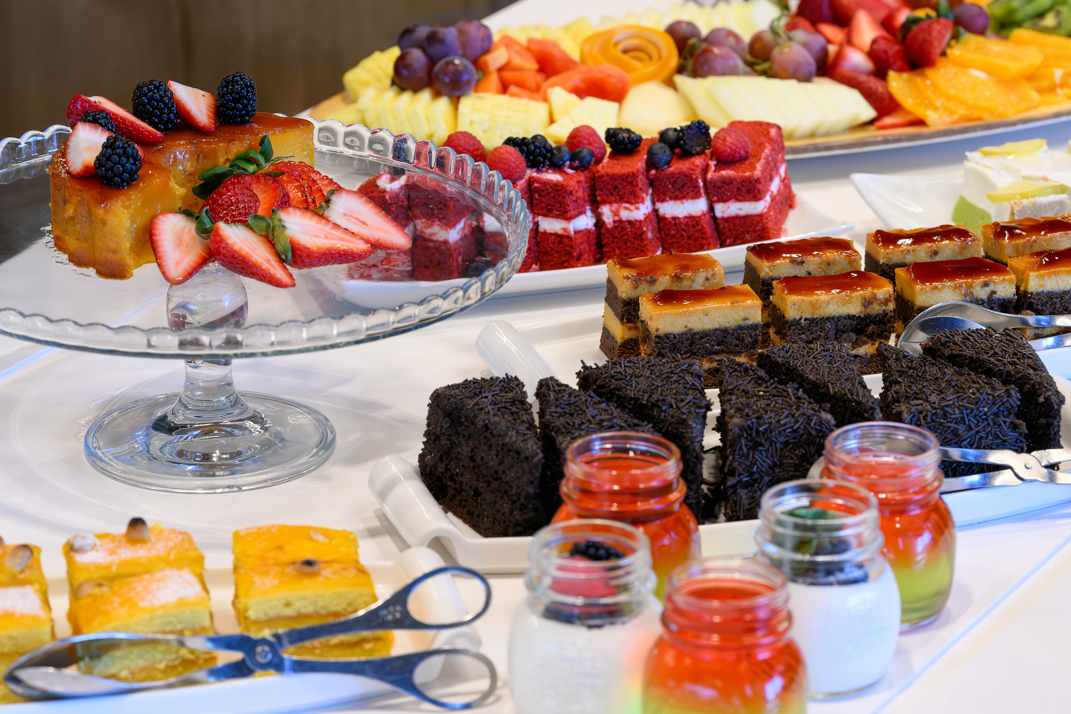 Dessert buffet with various cakes, pastries, flan, red velvet, and fruit platter.