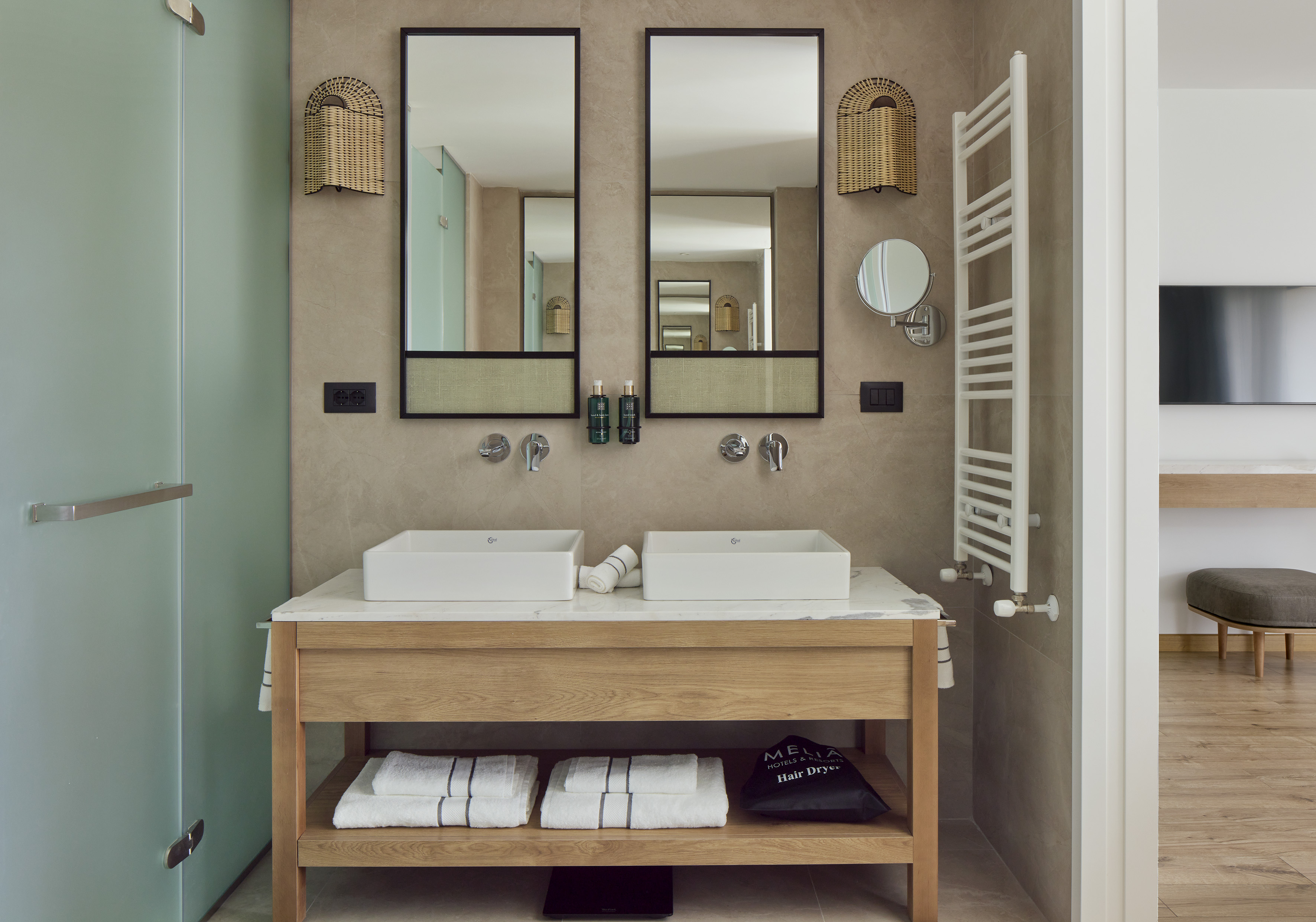 Modern hotel bathroom with double vanity, wood cabinet, and woven mirrors.