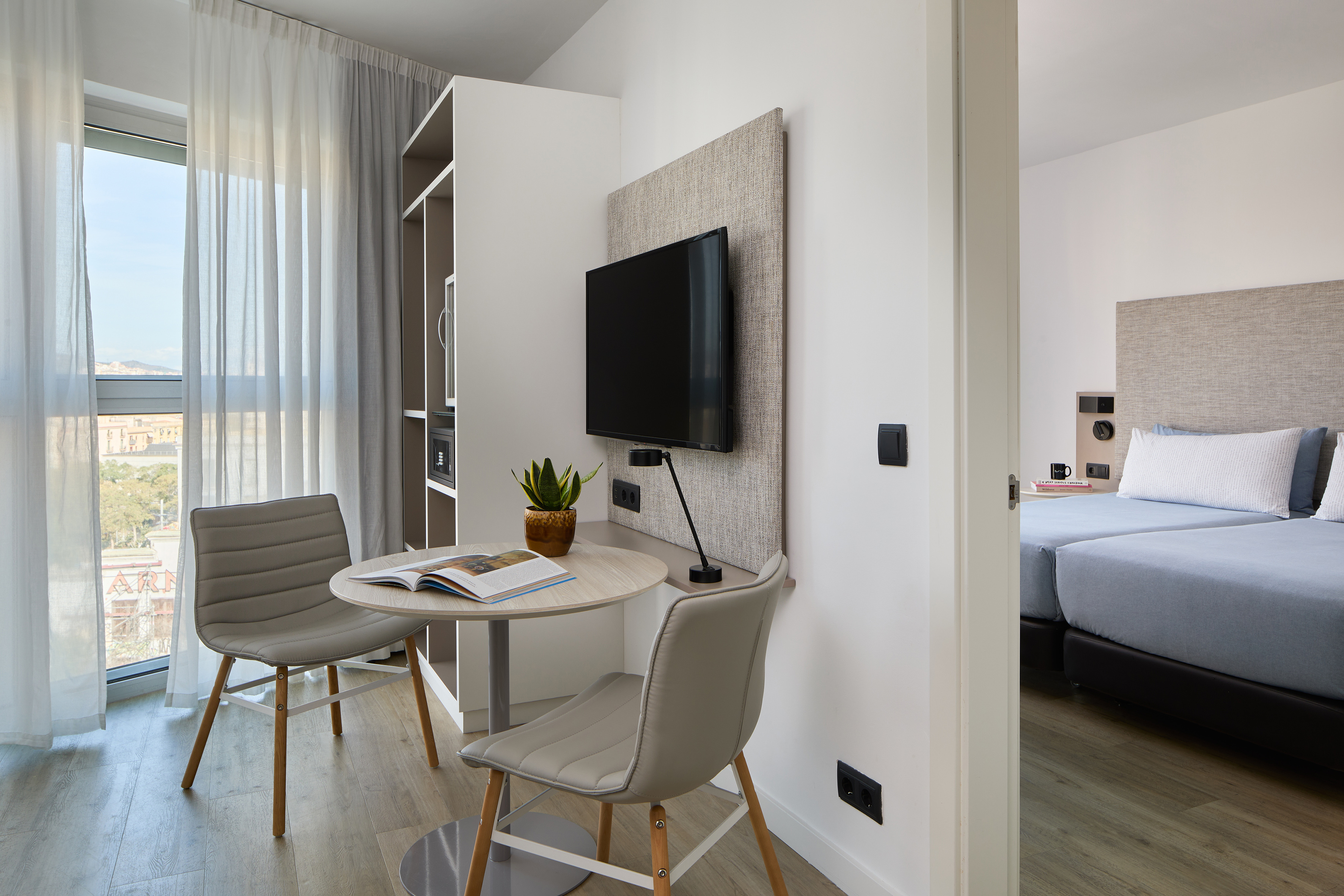 Modern hotel studio with dining area, wall-mounted TV, and adjacent bedroom.