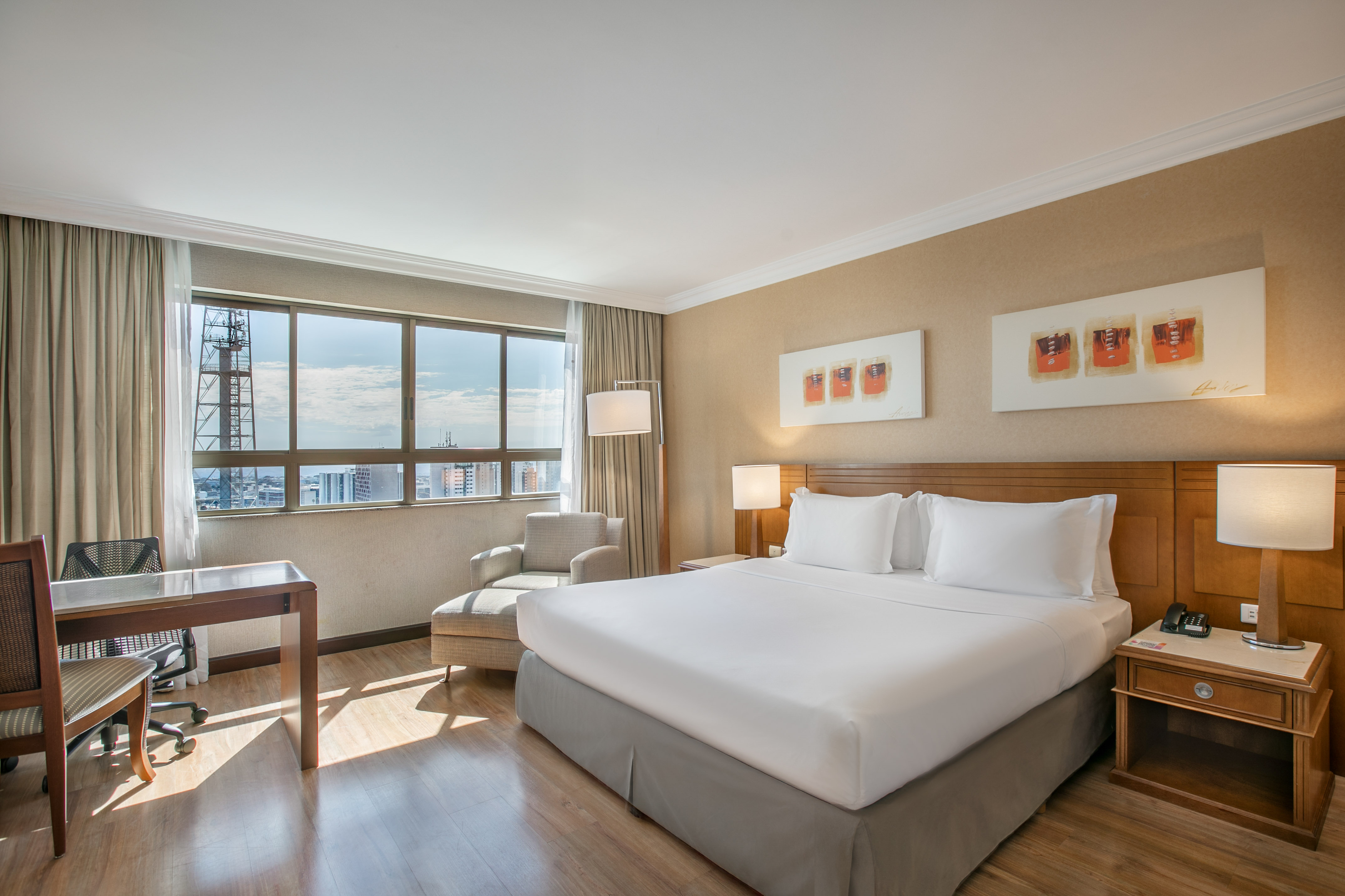 Hotel bedroom with king bed, desk, large window city view, and armchair.
