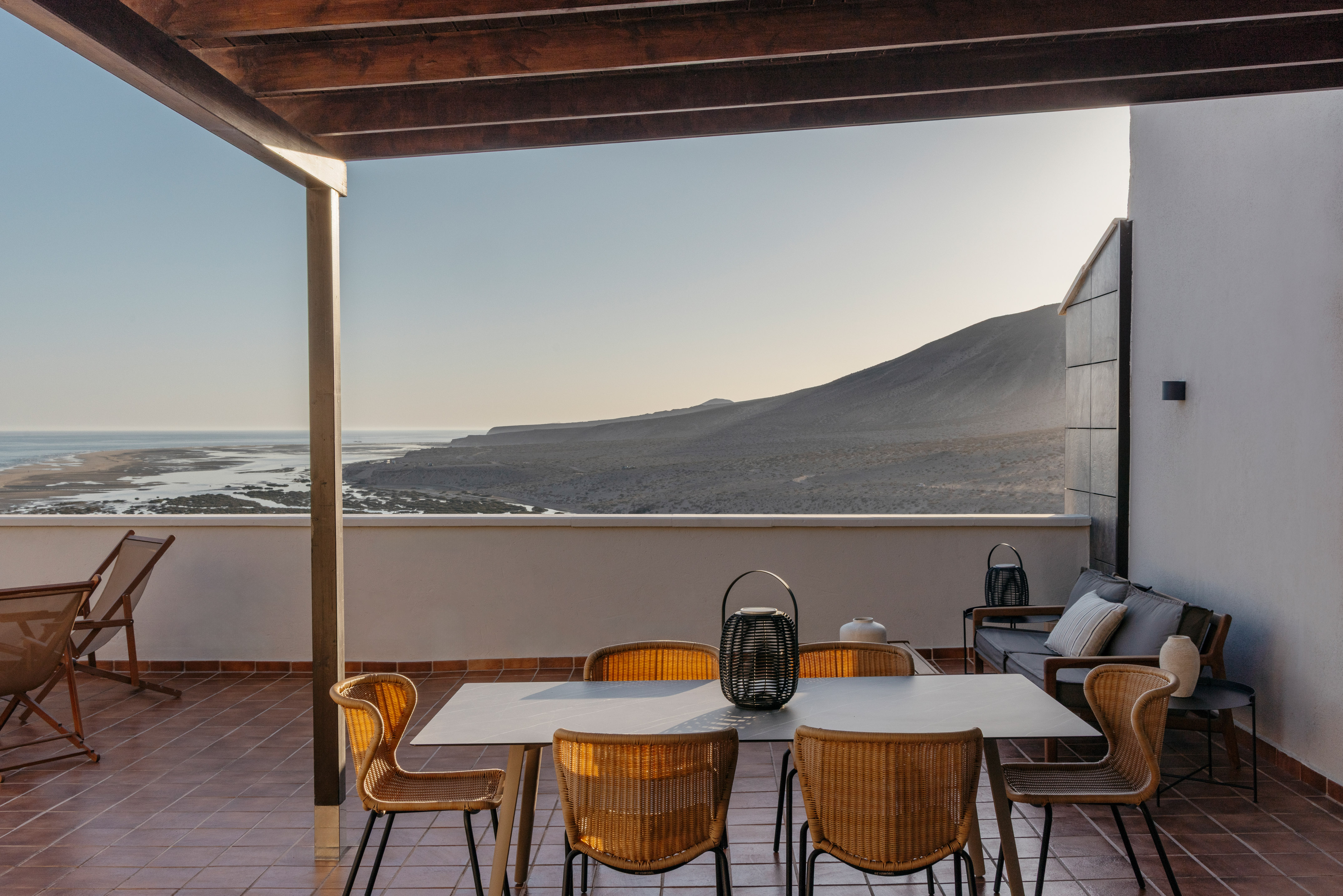 Coastal terrace dining with wicker chairs, ocean, and arid mountain panorama.