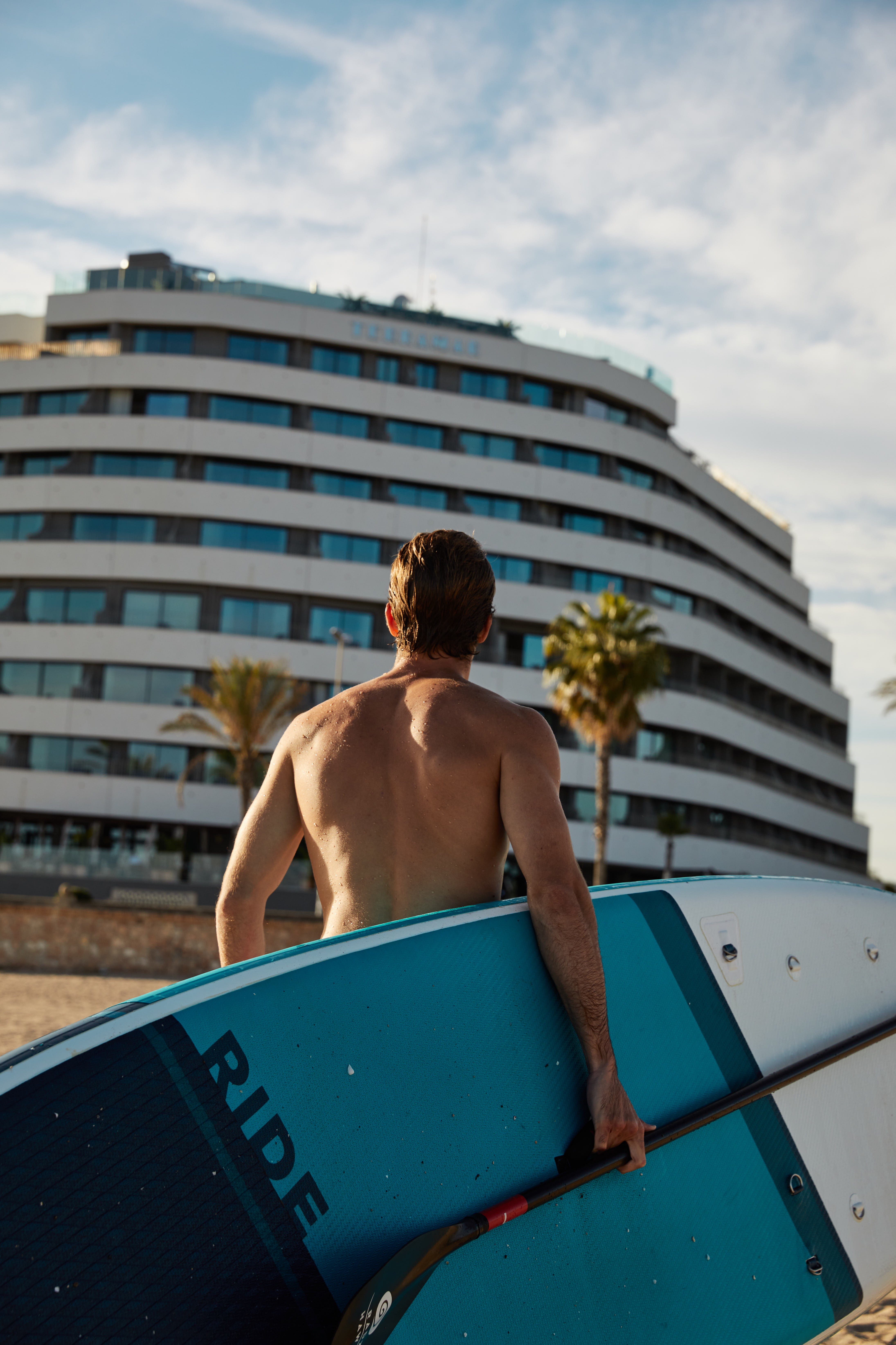 Man carrying blue SUP paddleboard on beach in front of Hotel Terramar.