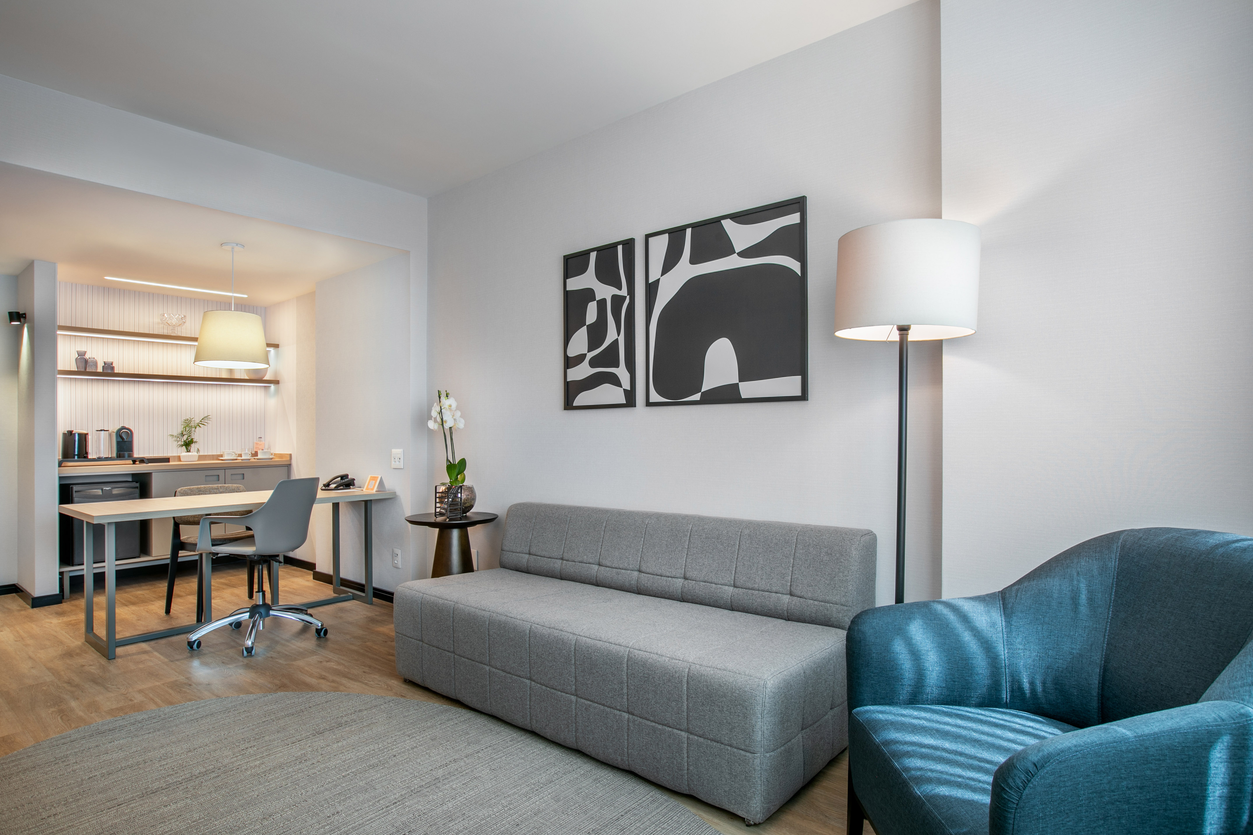 Modern hotel suite with gray sofa, blue armchair, desk, and kitchenette.