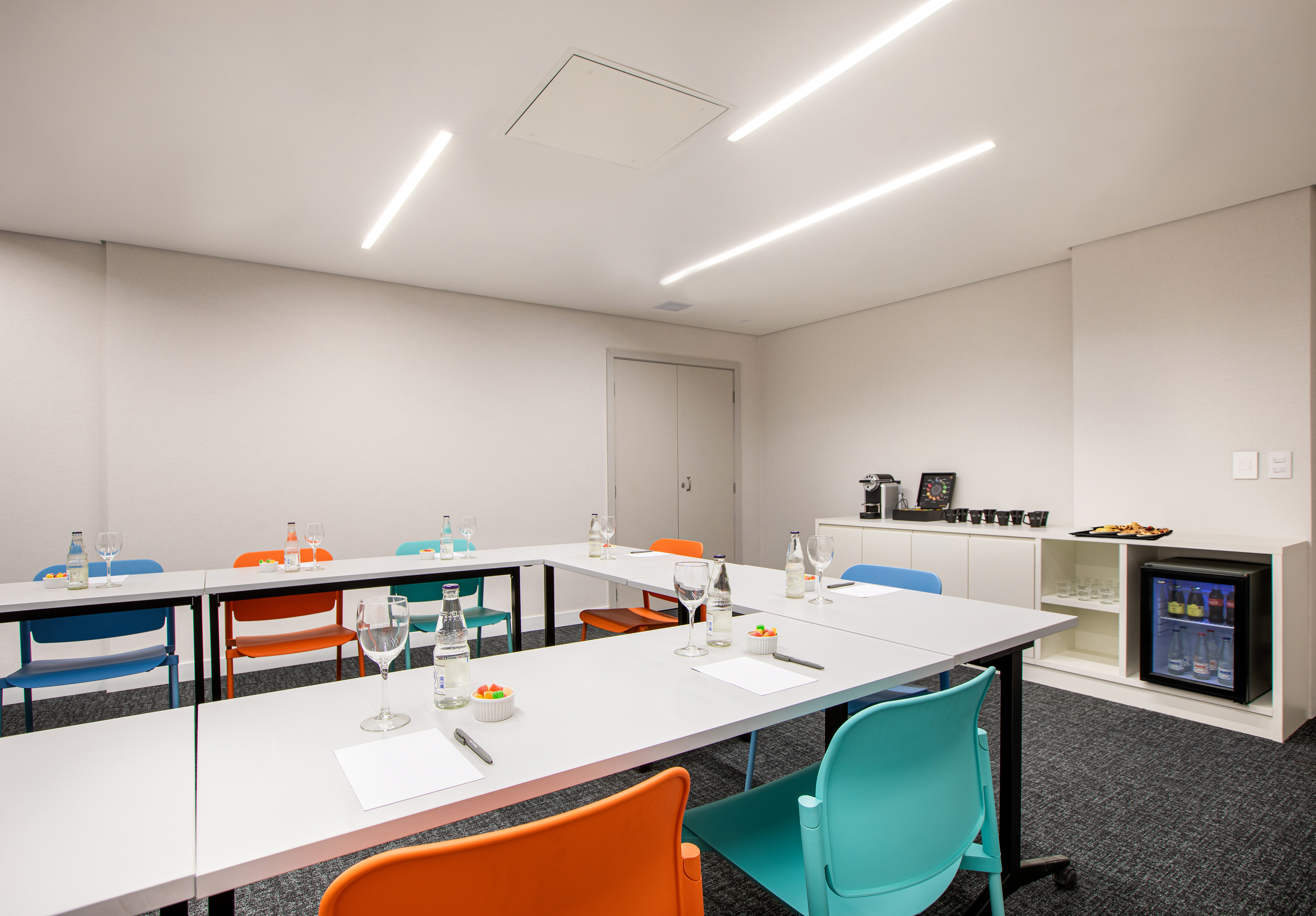 Modern conference room with U-shaped tables, vibrant chairs, and refreshment station.