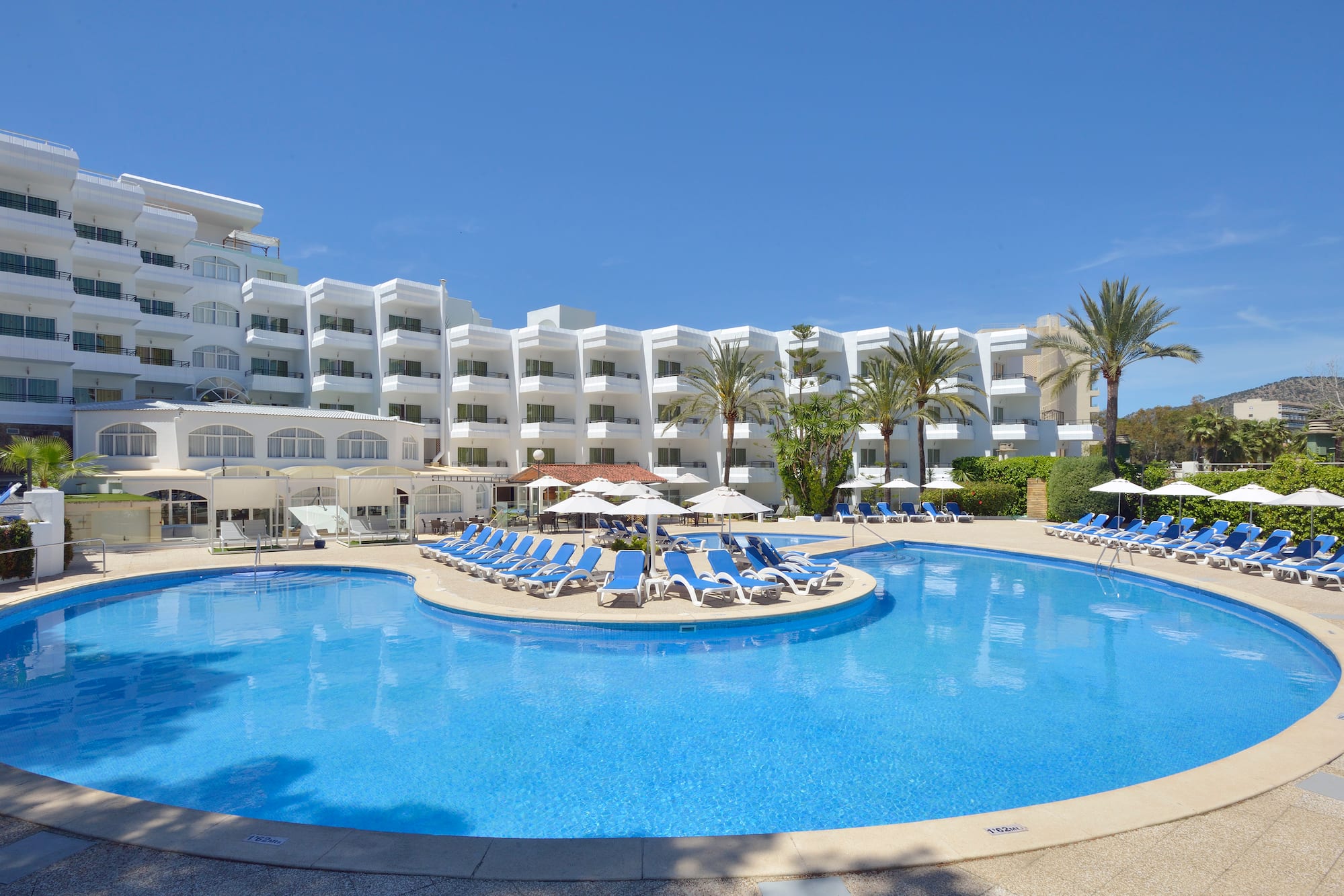 Sunny resort hotel with large outdoor pool, blue sun loungers, and palm trees.