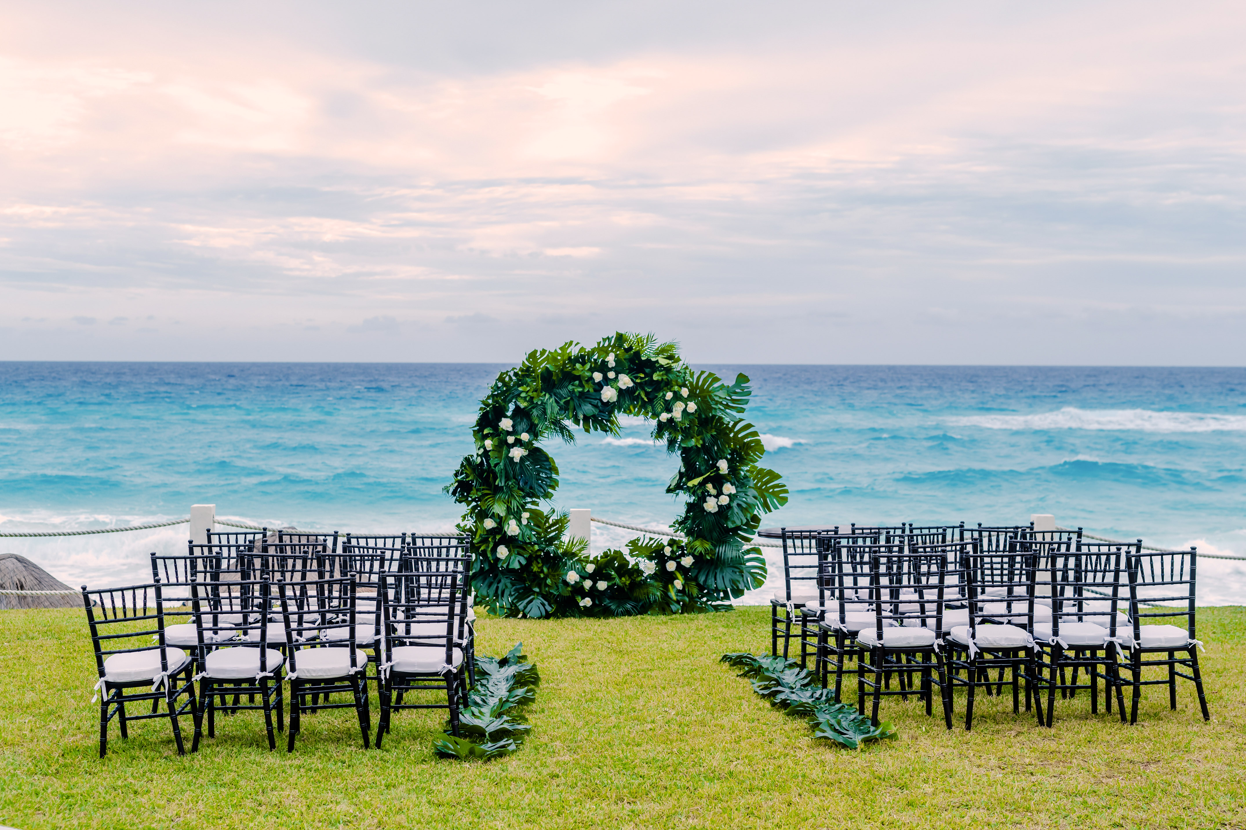 Tropical beach wedding ceremony with green arch, black chairs, and ocean view.