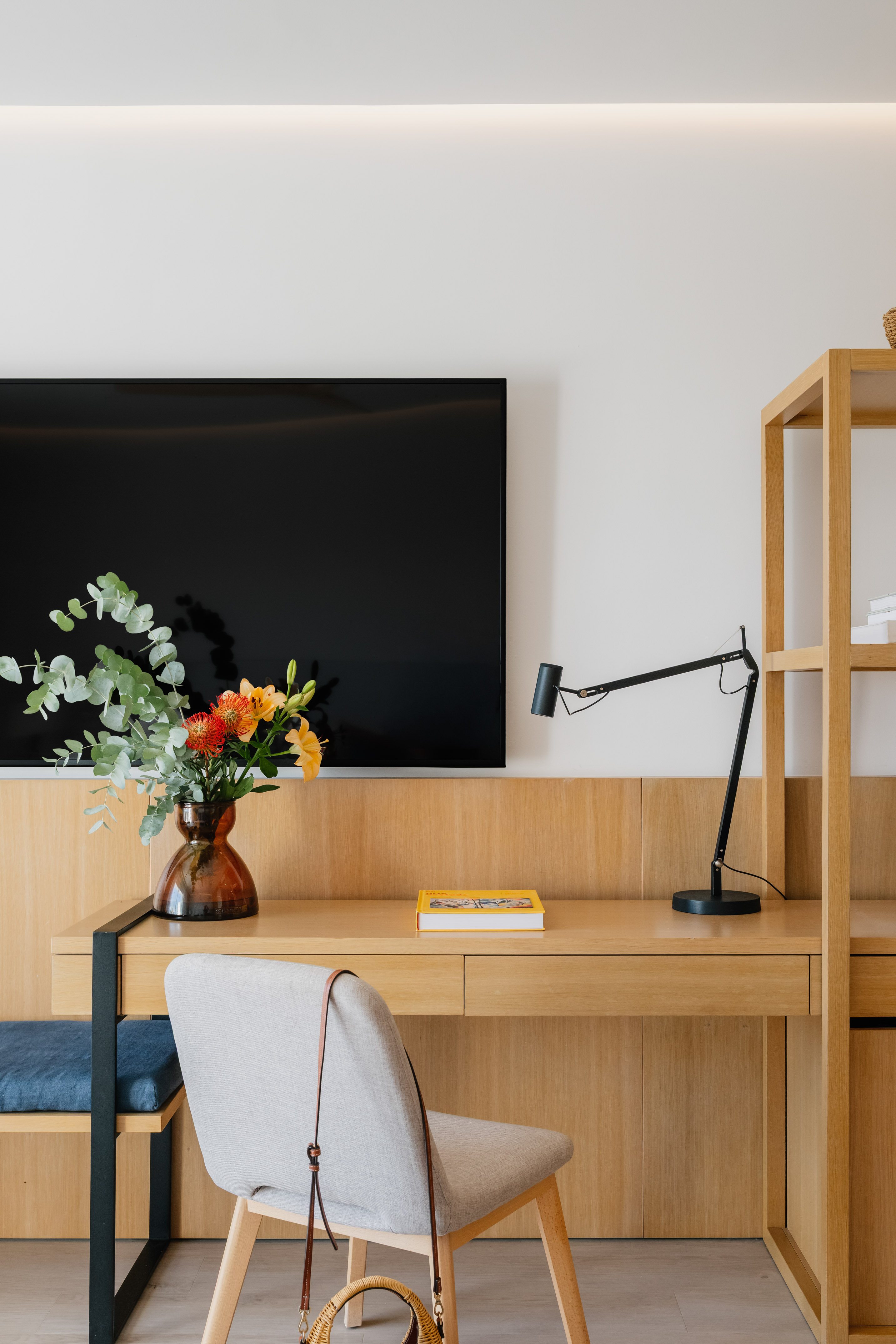 Minimalist home office with wooden desk, TV, floral vase, and sleek black lamp.