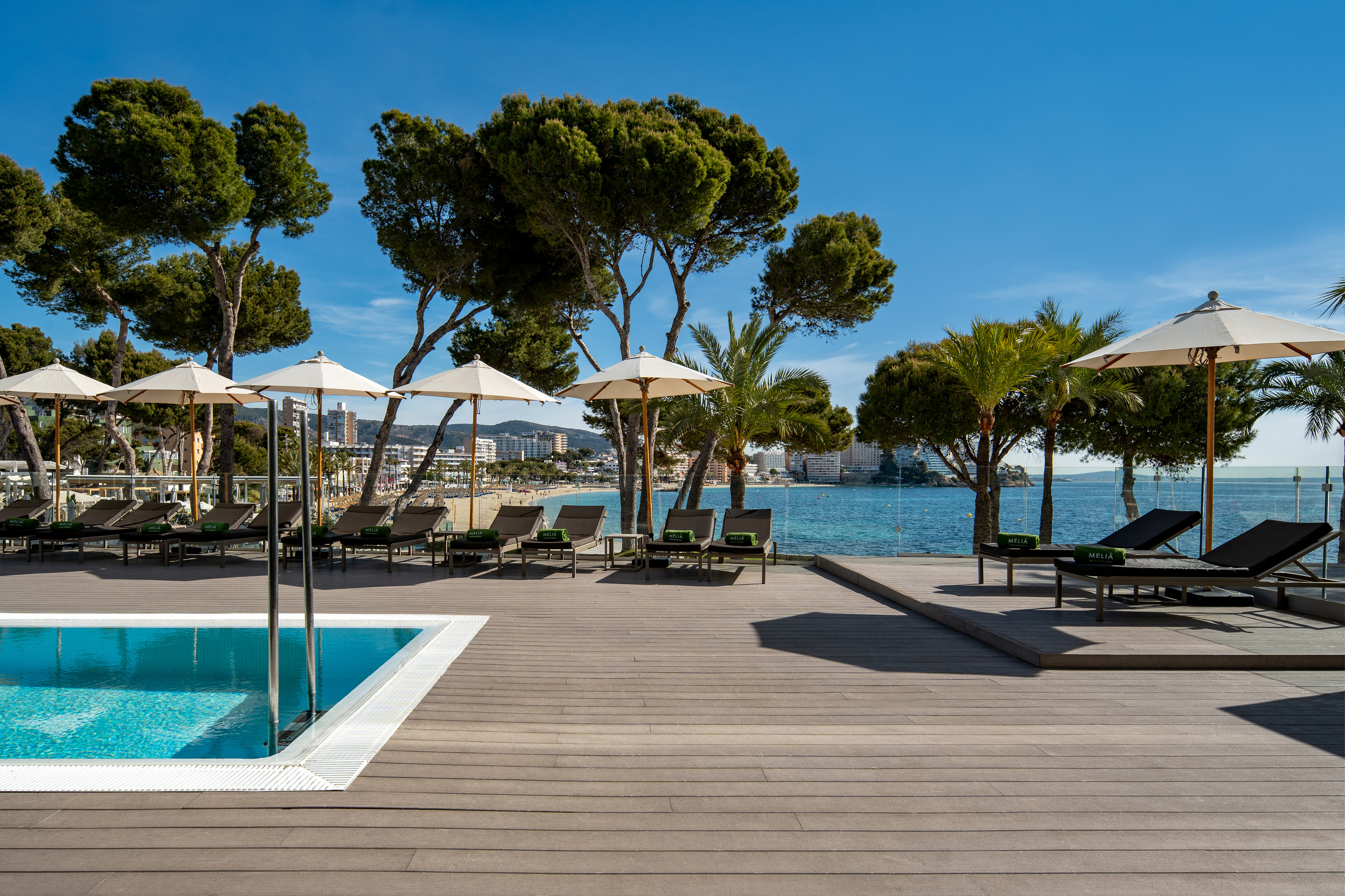 Poolside deck with sun loungers, umbrellas, clear blue sea, beach, and coastal trees.
