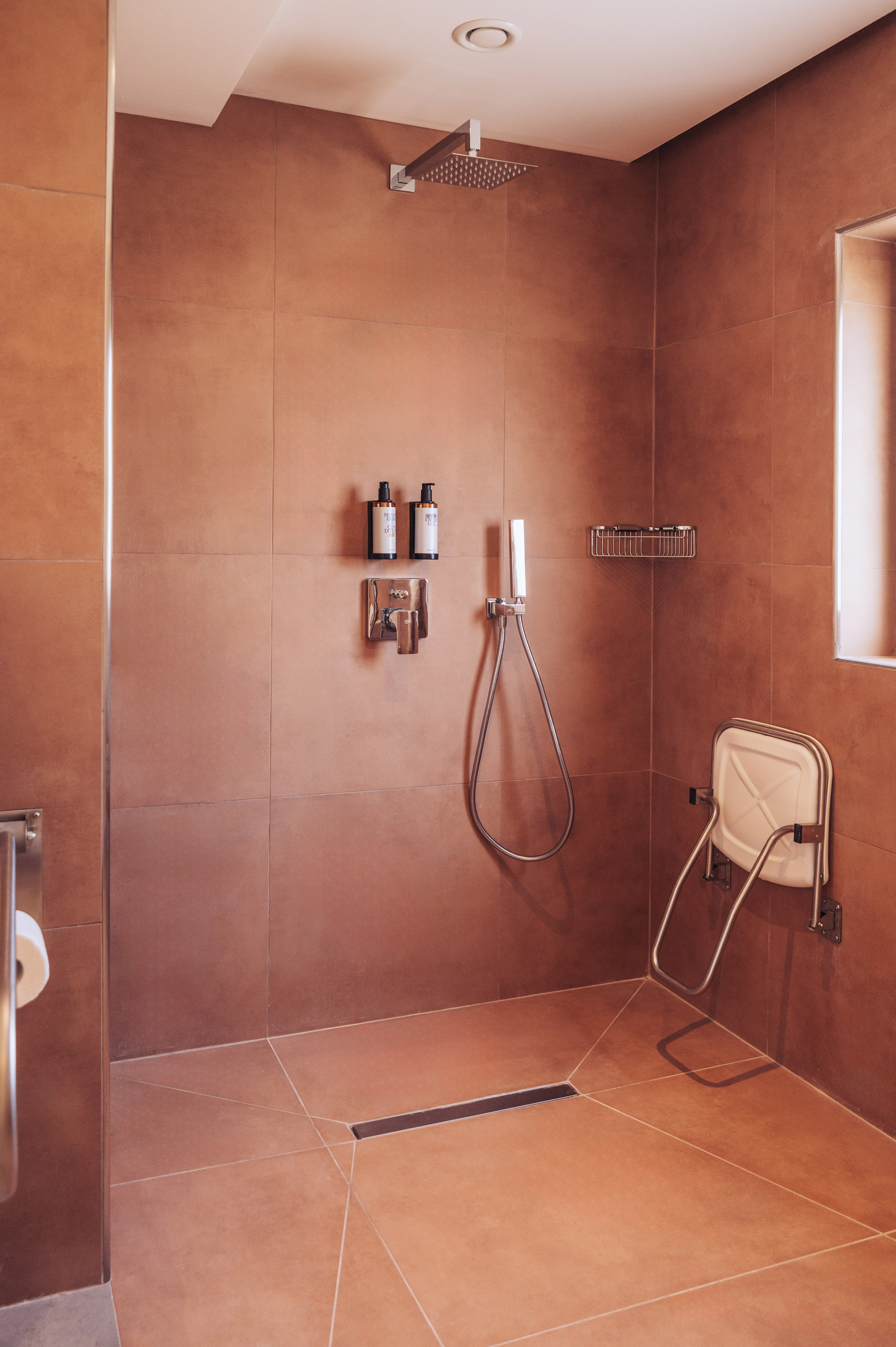 Accessible brown tiled shower with rain head, handheld, and folding seat.