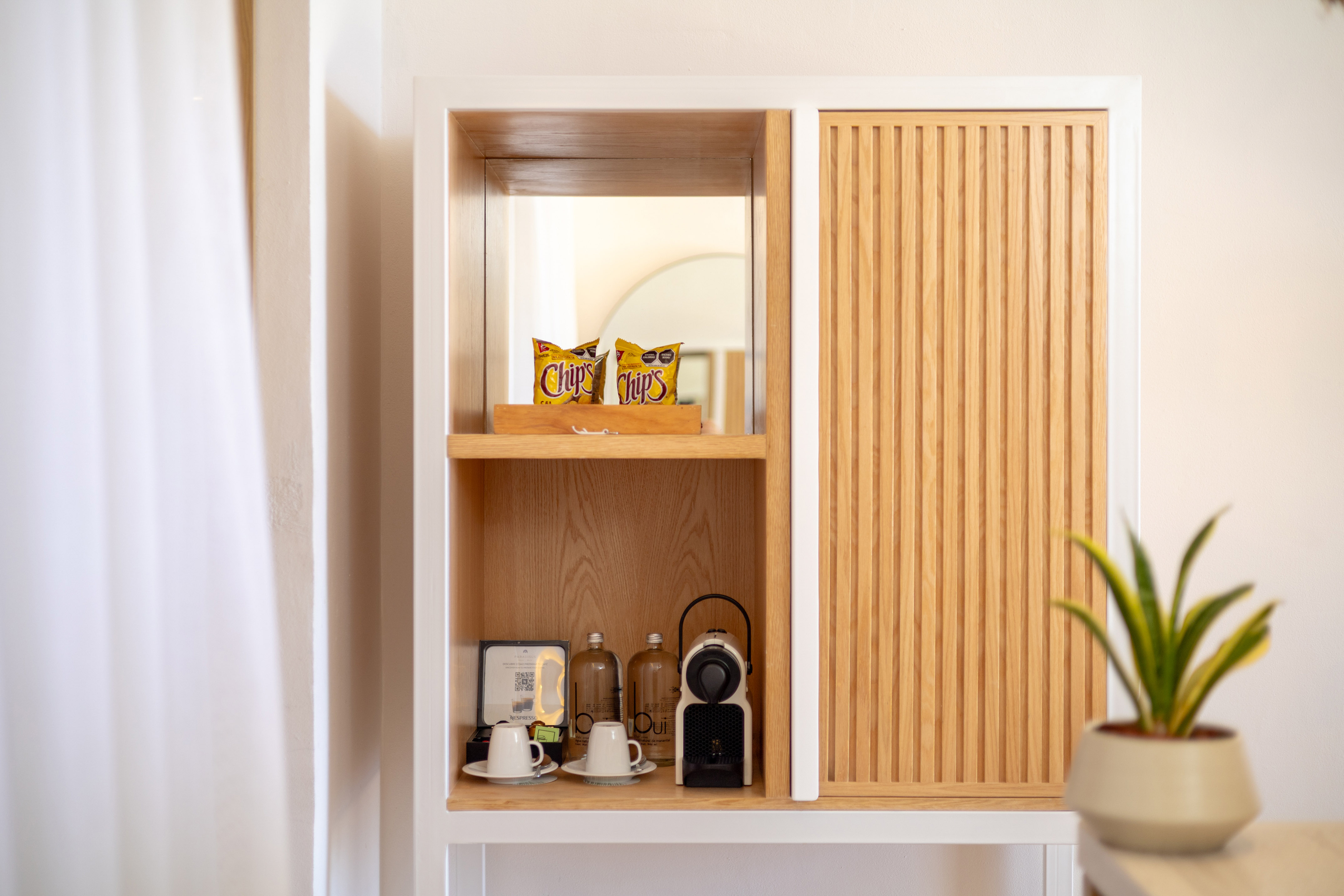 Modern wooden hotel minibar with Nespresso machine, snacks, and water bottles.