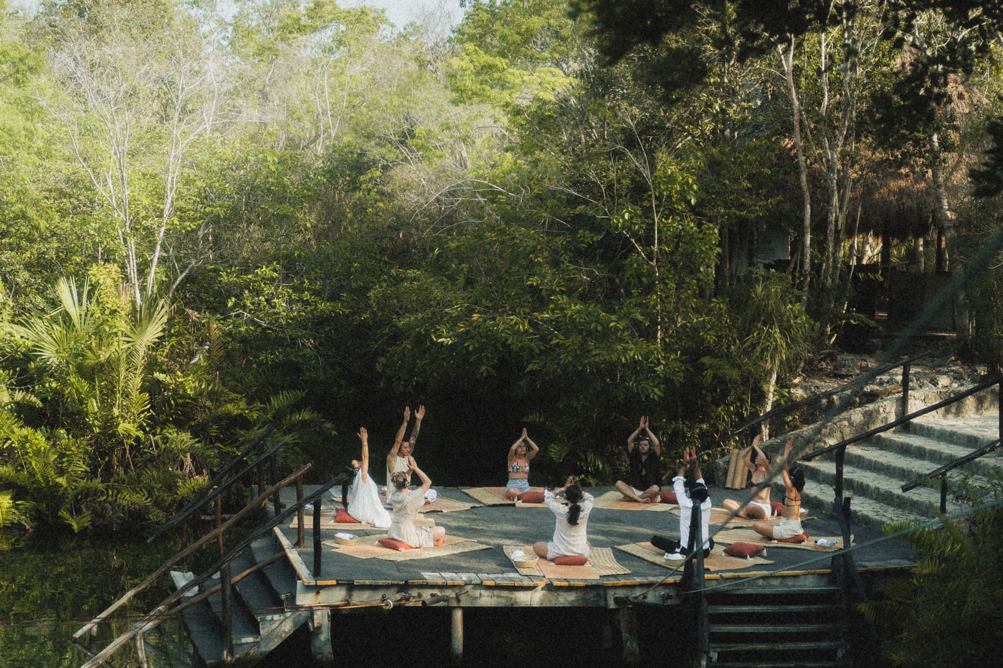 Yoga y meditación grupal en plataforma sobre agua de cenote, rodeado de selva.